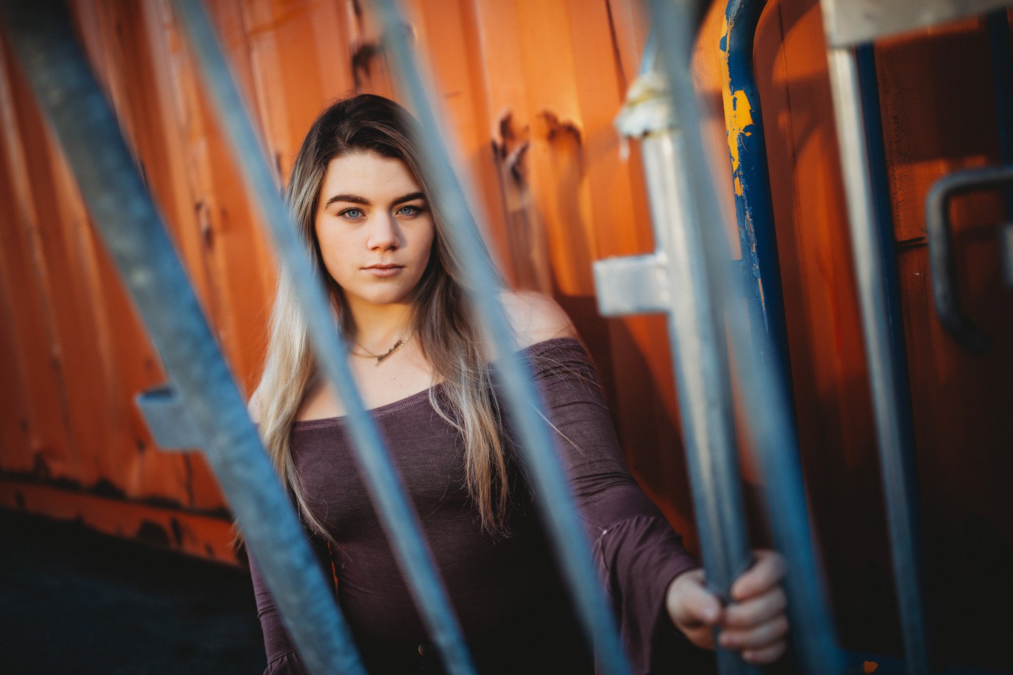 A young woman with long, blonde and brown hair and blue eyes standing behind metal bars, with an orange wooden wall in the background. She is wearing a dark burgundy off-shoulder top and looking confidently at the camera.