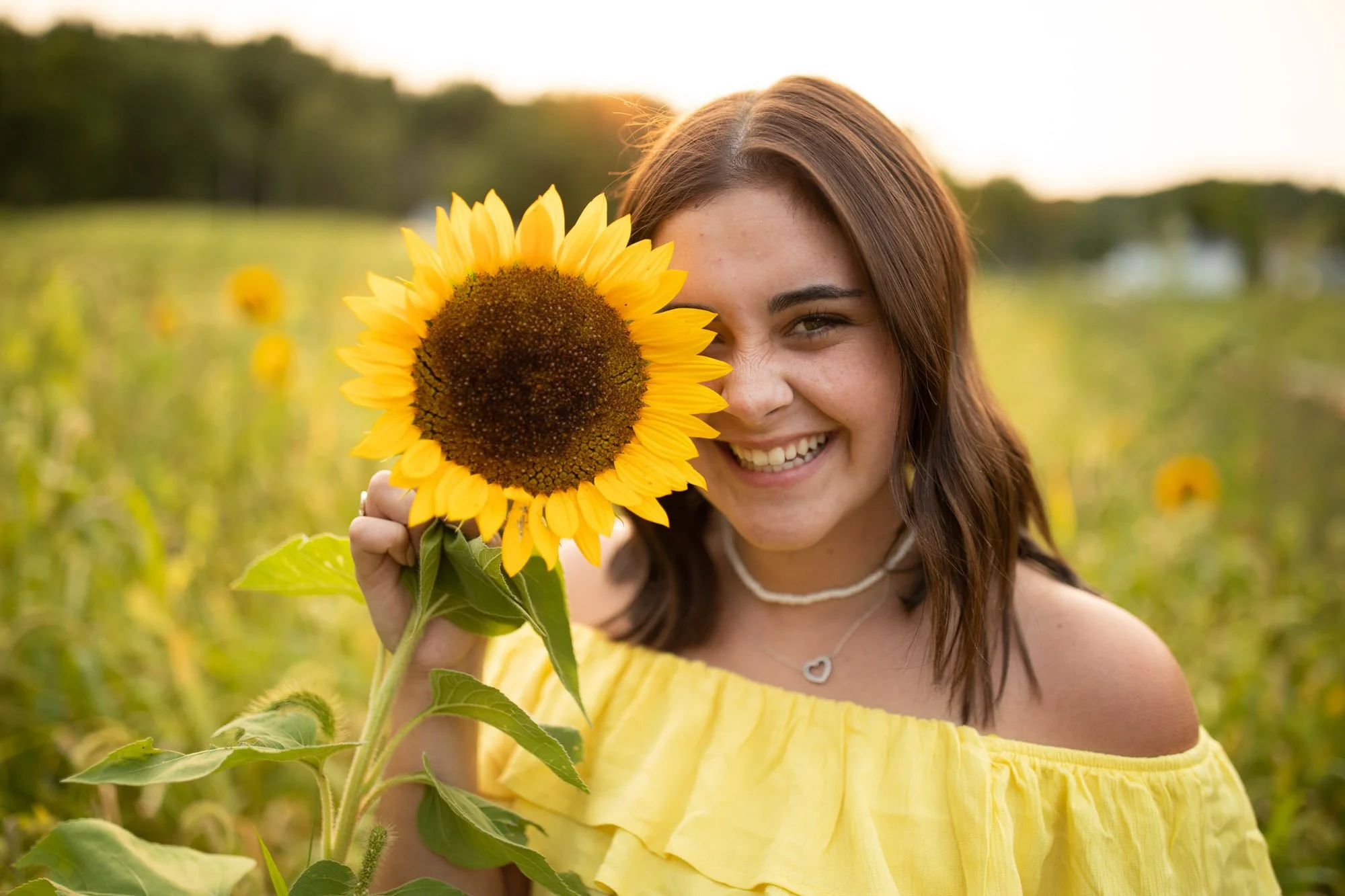 A young woman with brown hair and a yellow off-shoulder top holding a sunflower, smiling in a sunflower field at sunset.