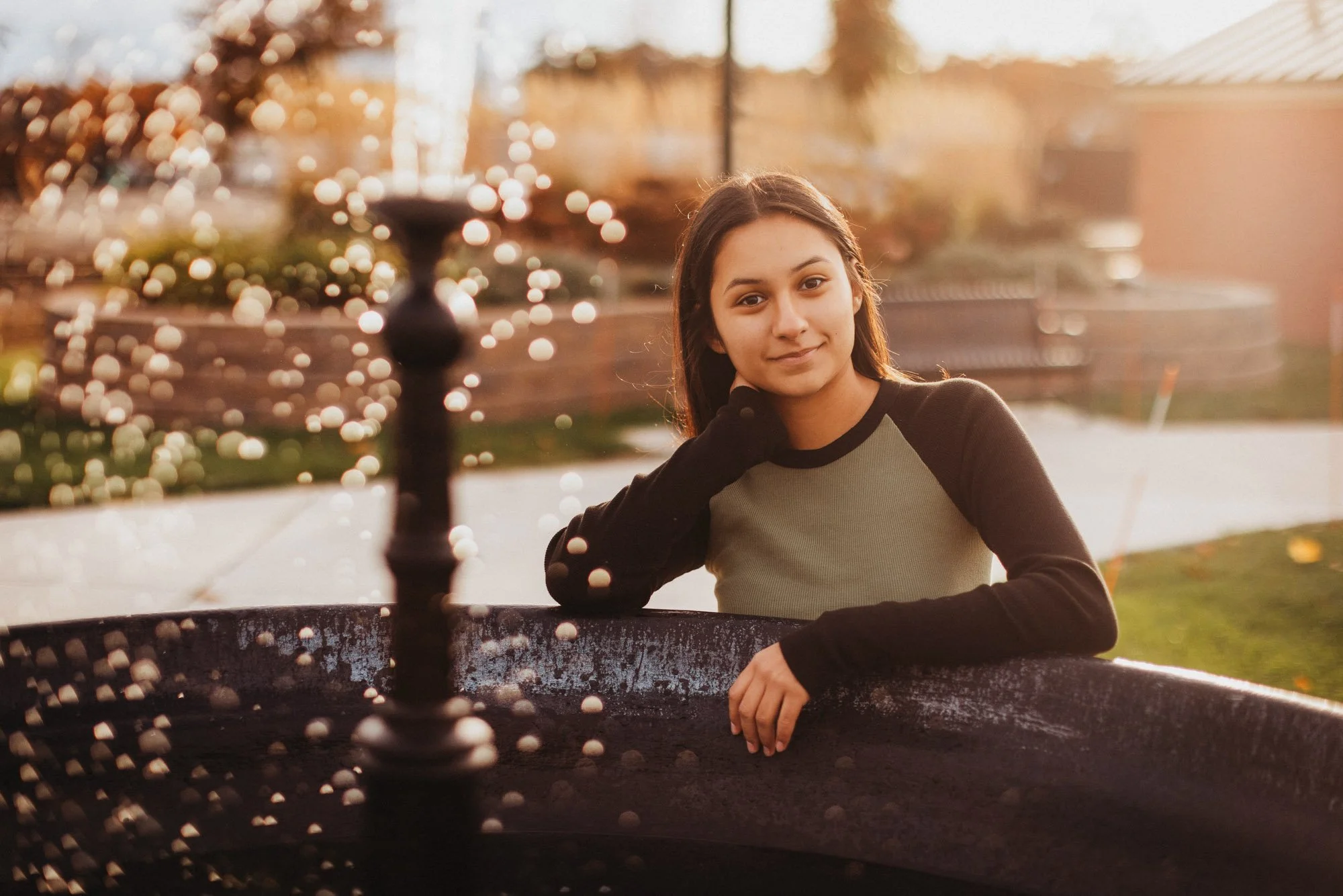 A young woman with long brown hair sits outdoors on a sunny day, resting her chin on her hand, with a relaxed expression. She is wearing a black and green long-sleeve shirt and is near a large, round black fire pit with a blurred background of trees 