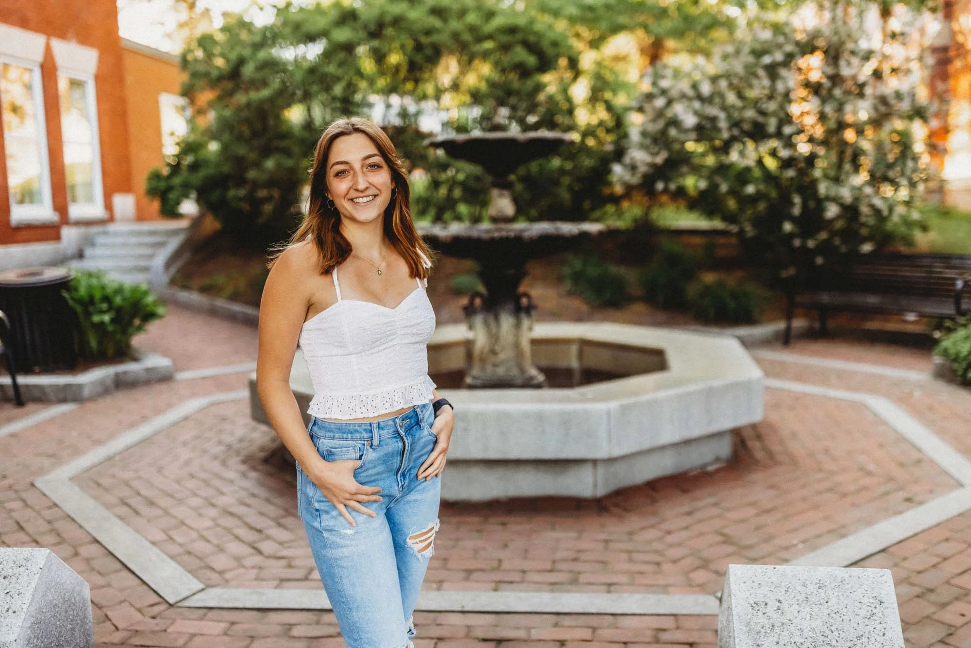 A young woman with shoulder-length brown hair, wearing a white sleeveless top and ripped blue jeans, smiling and standing in front of a fountain in a brick-paved courtyard with greenery and benches.