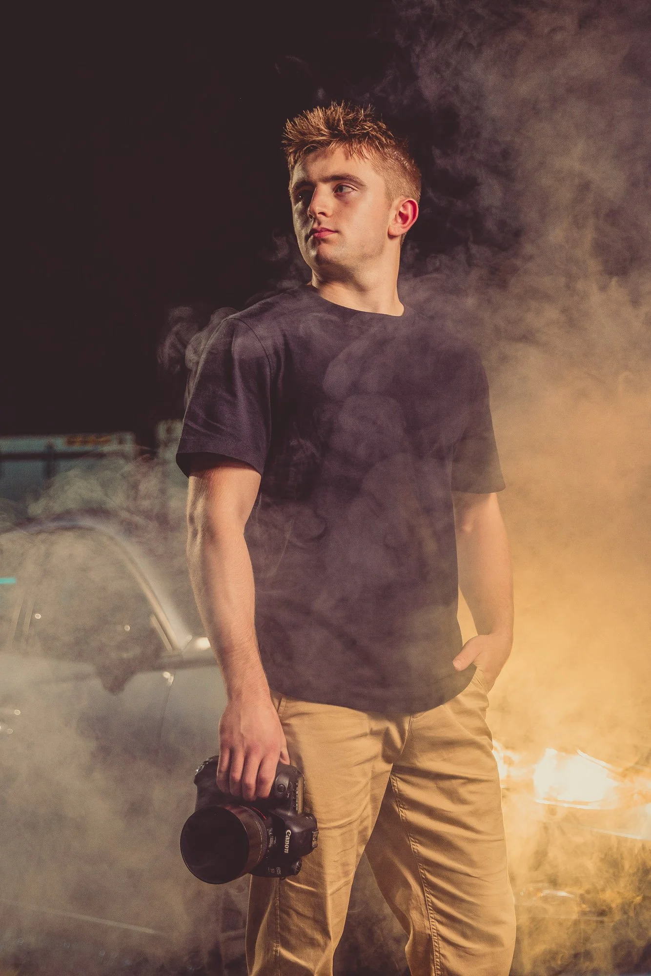 Young man standing with a camera in hand, surrounded by smoke and lit by warm light, at night