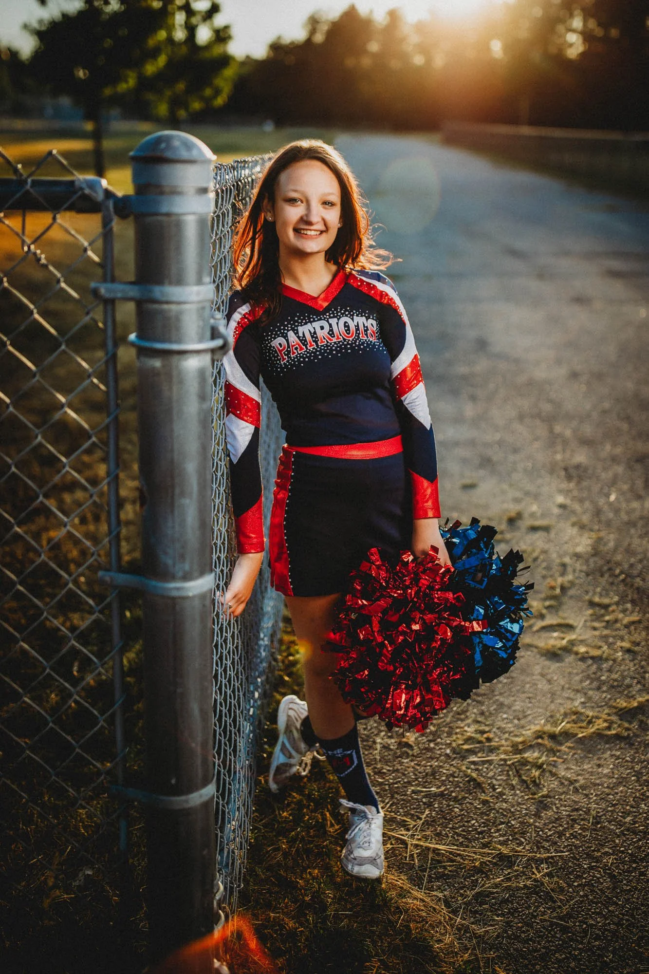 Young female cheerleader in Patriots uniform holding pom-poms, standing next to a chain-link fence during sunset at a sports field.
