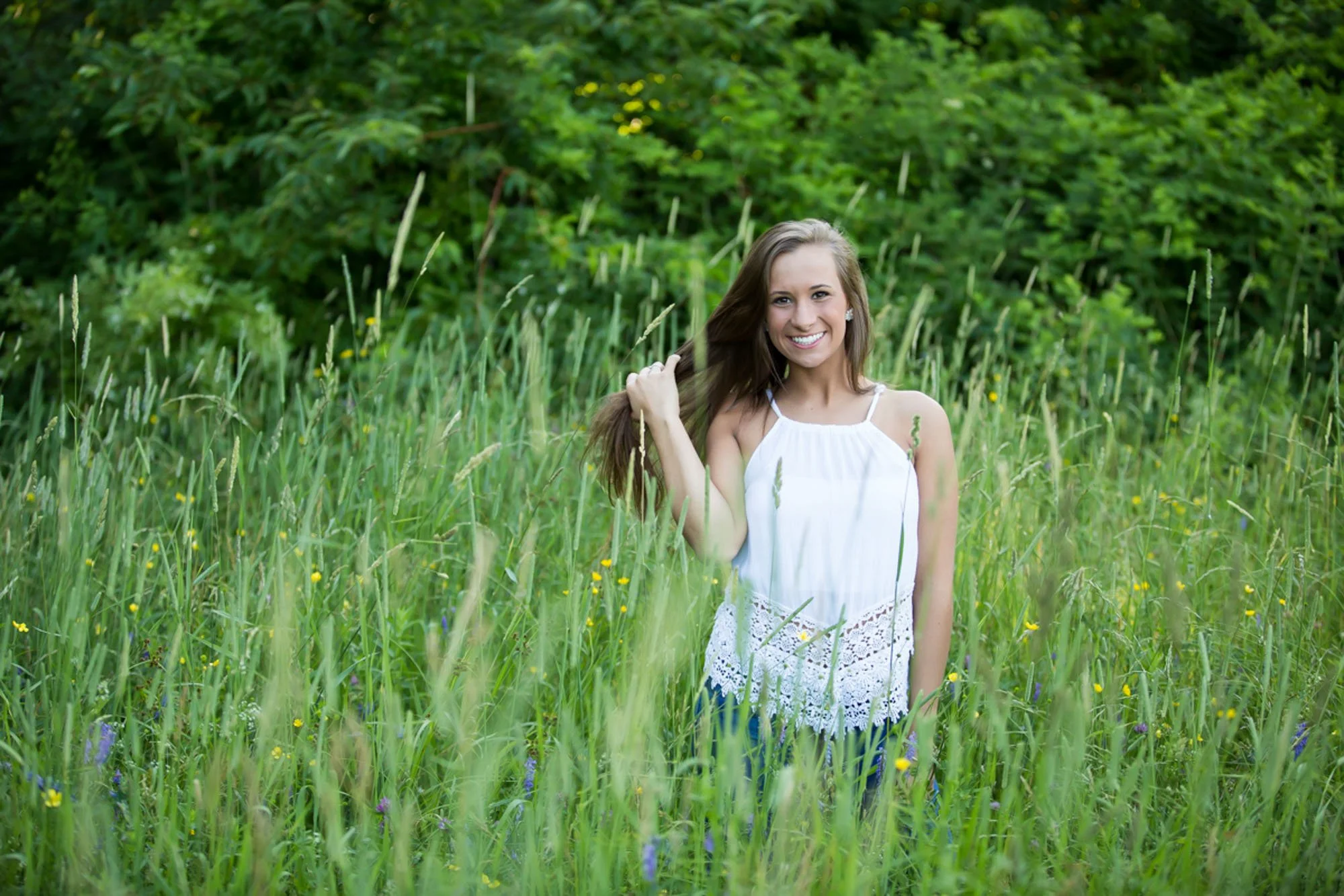 A young woman in a white top smiling while standing in a grassy meadow surrounded by tall green grass and wildflowers.