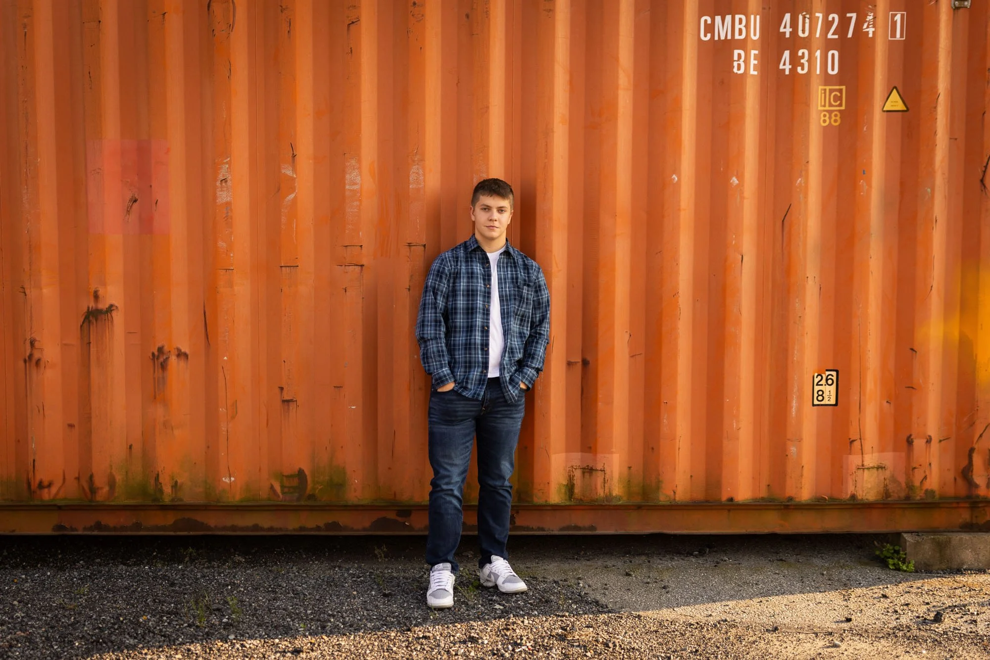 A young man wearing a blue plaid shirt, jeans, and white sneakers standing in front of an orange shipping container with markings and moss at the bottom, outdoors on a gravel surface.