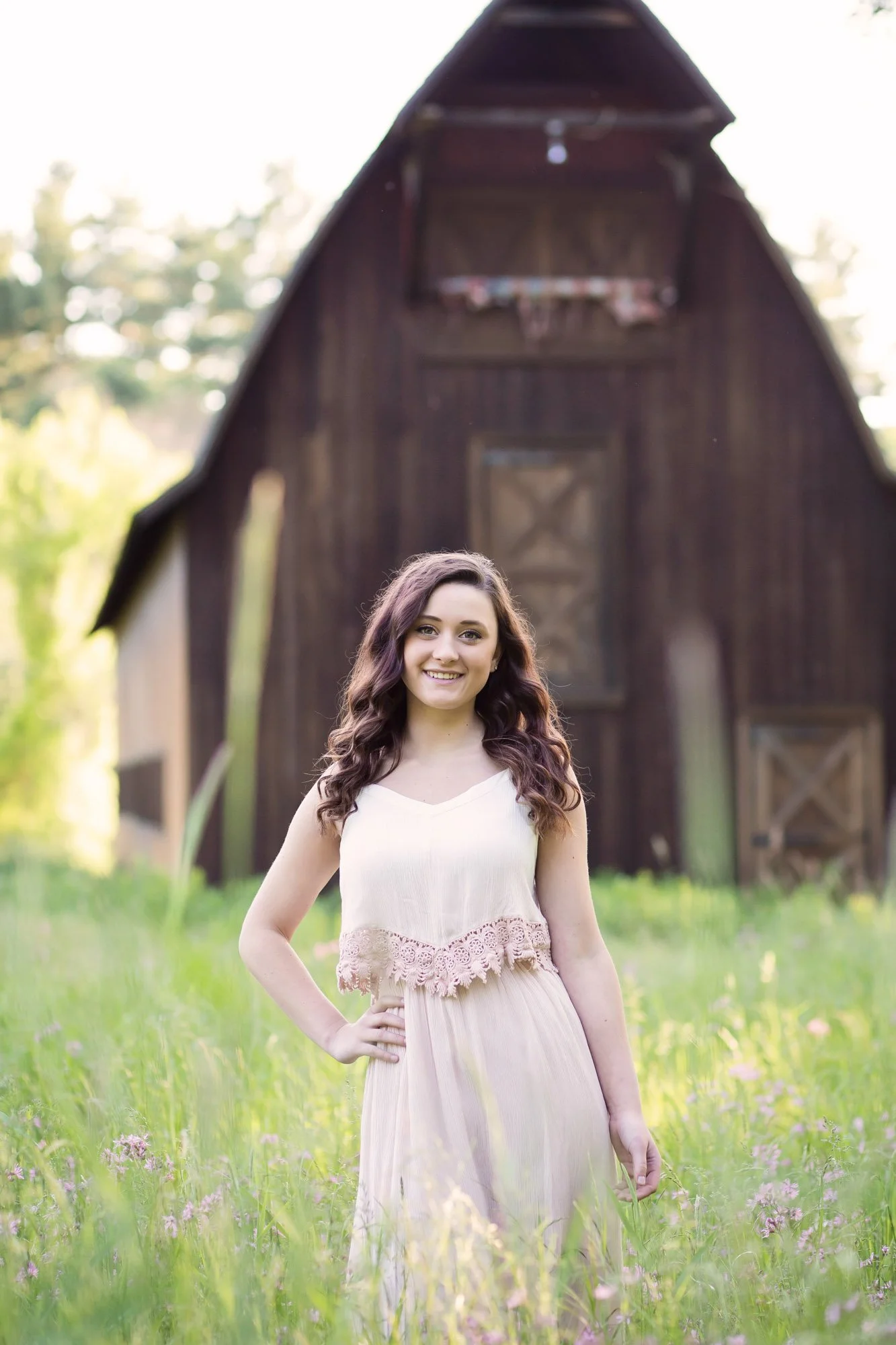 A young woman with wavy brown hair wearing a cream-colored dress with lace details, standing in a grassy field with pink flowers, smiling at the camera, with a large wooden barn in the background.
