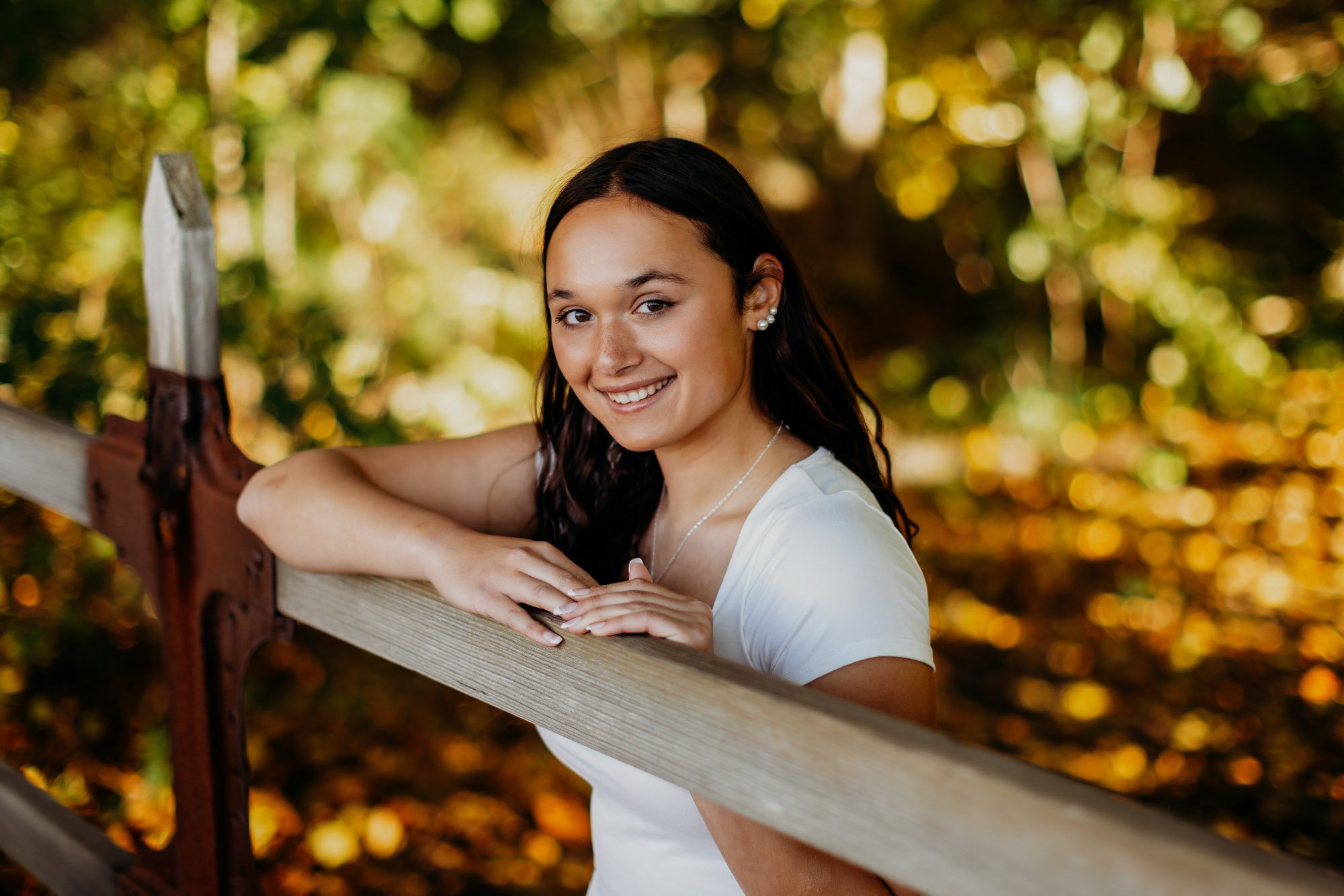 A young woman with dark curly hair, wearing a white shirt and pearl earrings, smiling and leaning on a wooden fence outdoors during autumn with yellow and orange leaves in the background.