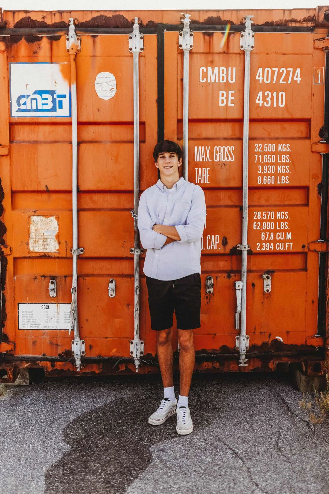 A young man with dark hair, wearing a white long-sleeve shirt, black shorts, white socks, and white sneakers, standing with arms crossed in front of an orange shipping container with white and blue markings.