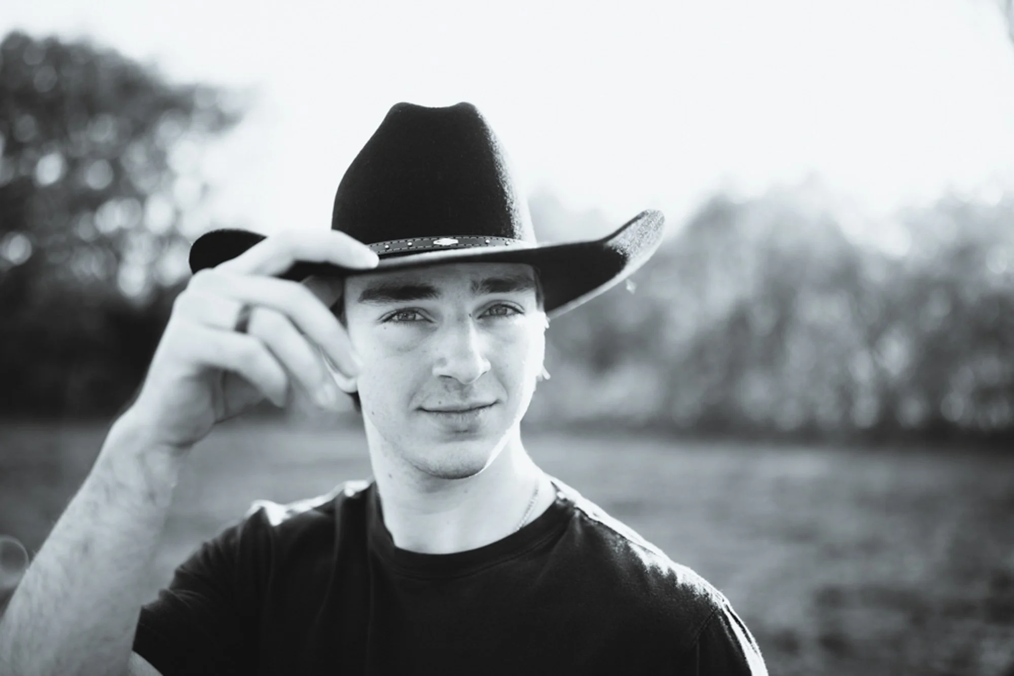A young man wearing a cowboy hat and a black t-shirt, holding the brim of his hat with his right hand, outdoors with blurred trees in the background, in black and white.