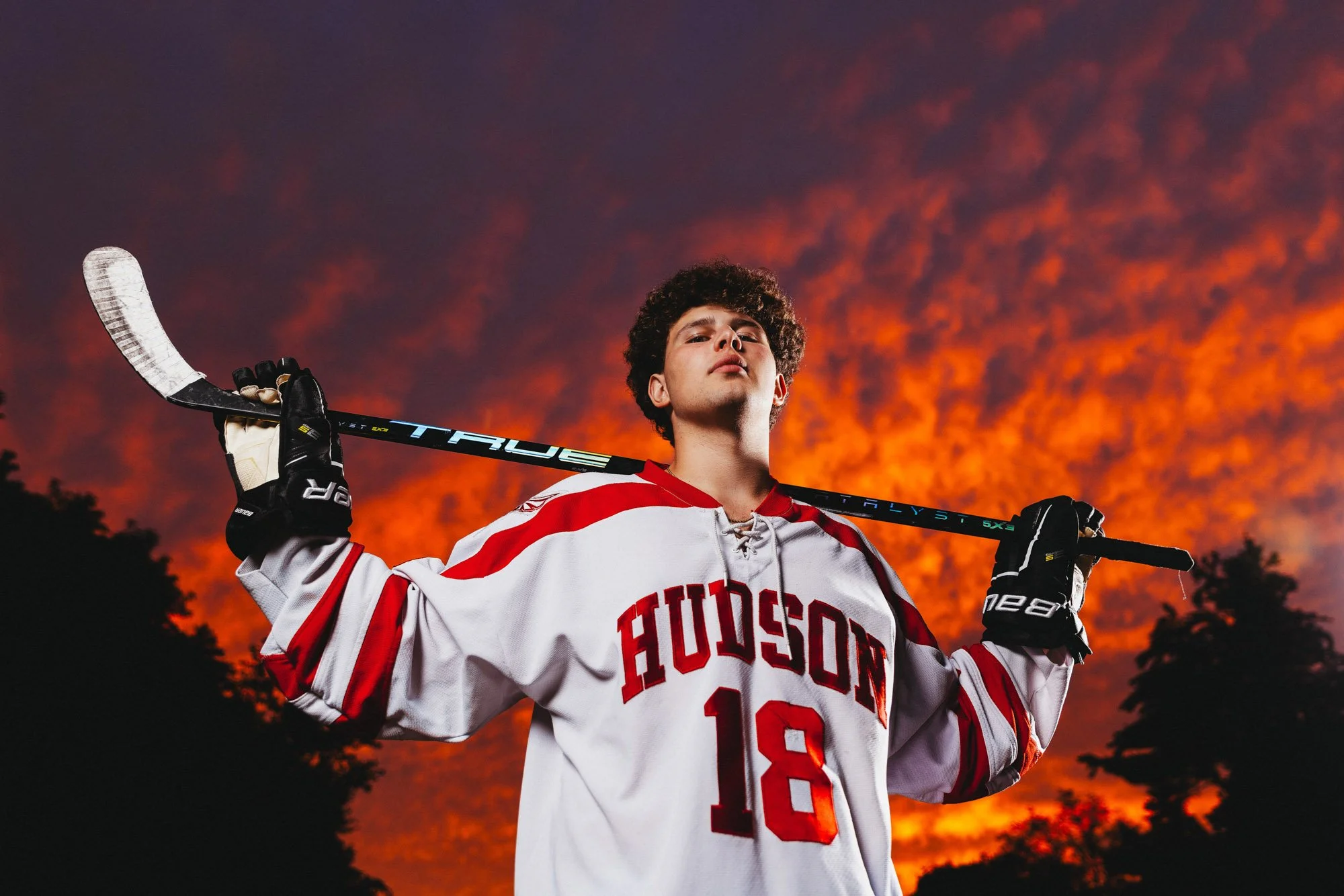 Hockey player in a white and red jersey with the number 18, standing against a dramatic orange and purple sunset sky, holding a hockey stick over his shoulders.