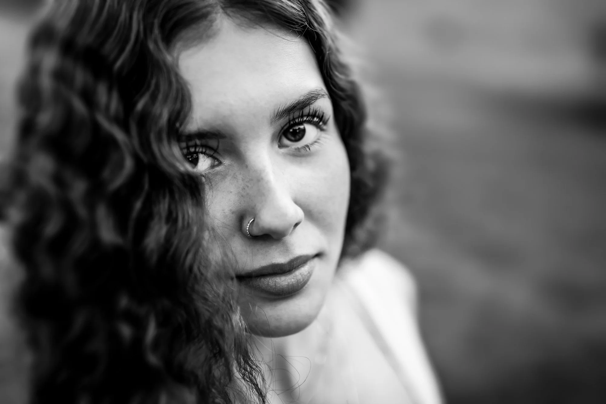 Black and white close-up portrait of a young woman with curly hair, a nose ring, and expressive eyes looking at the camera.