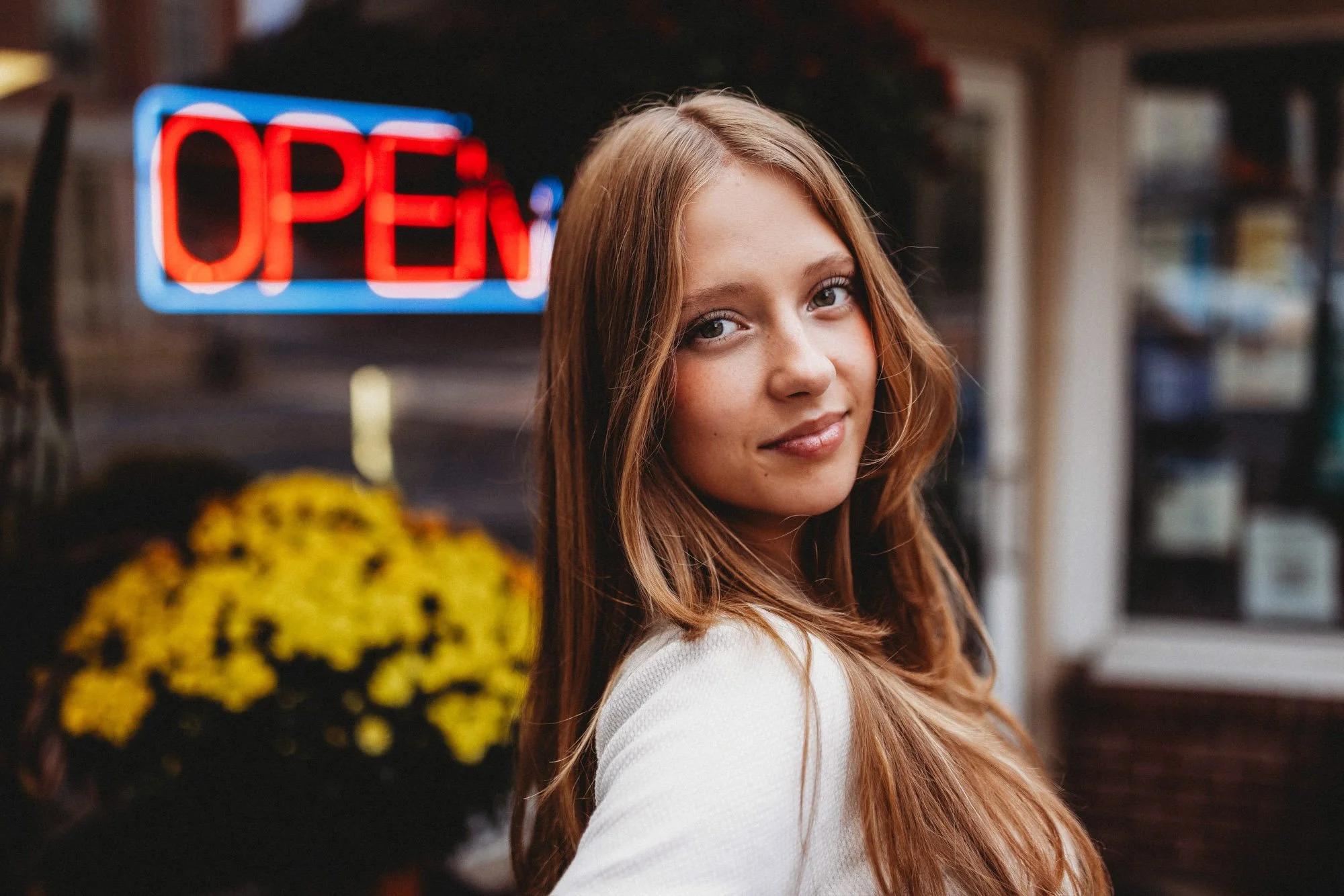 A young woman with long, wavy light brown hair and fair skin standing outside near a store with a neon "OPEN" sign in the background and a pot of yellow flowers.