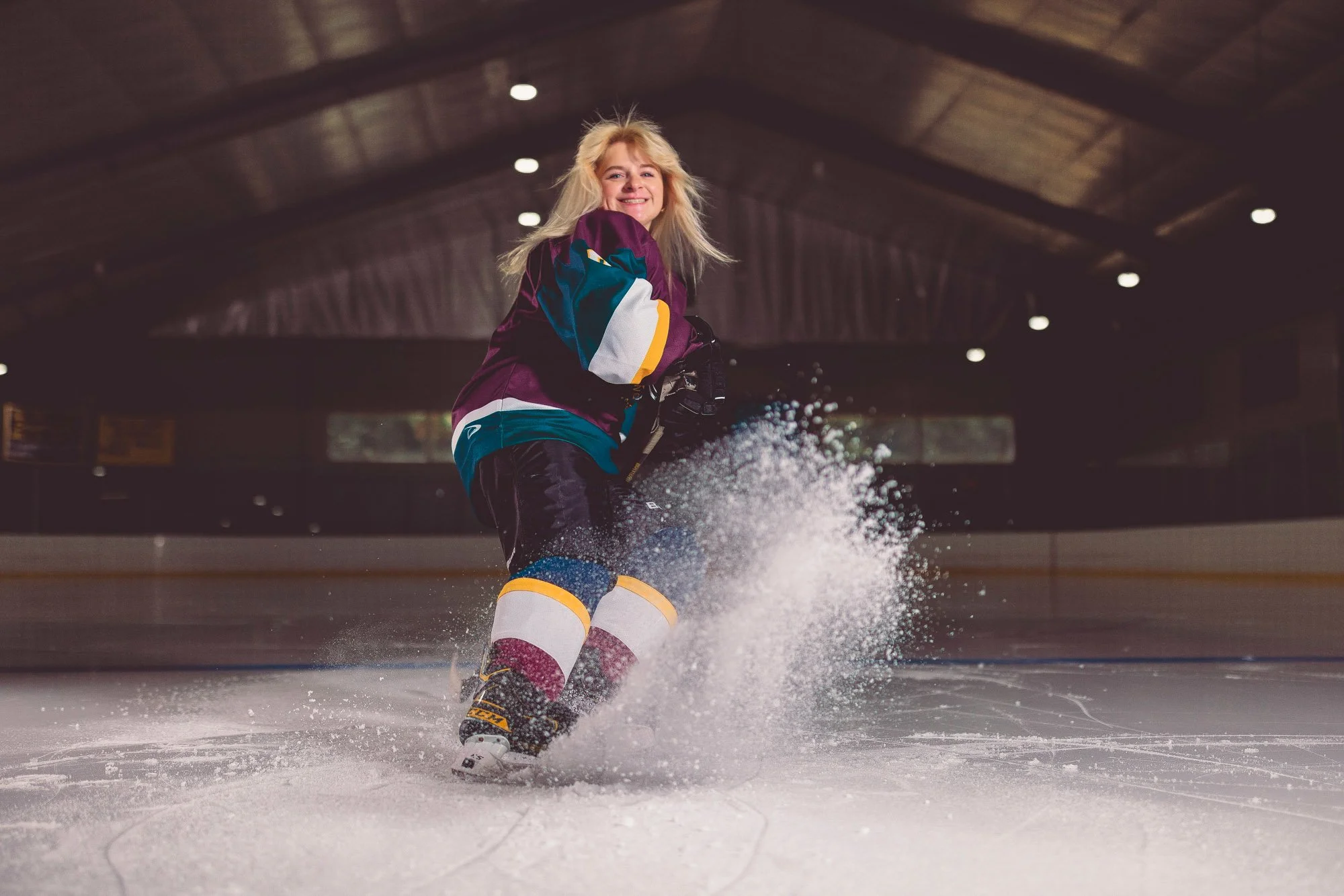 Person in colorful hockey gear smiling on an ice rink, skating and kicking up snow.