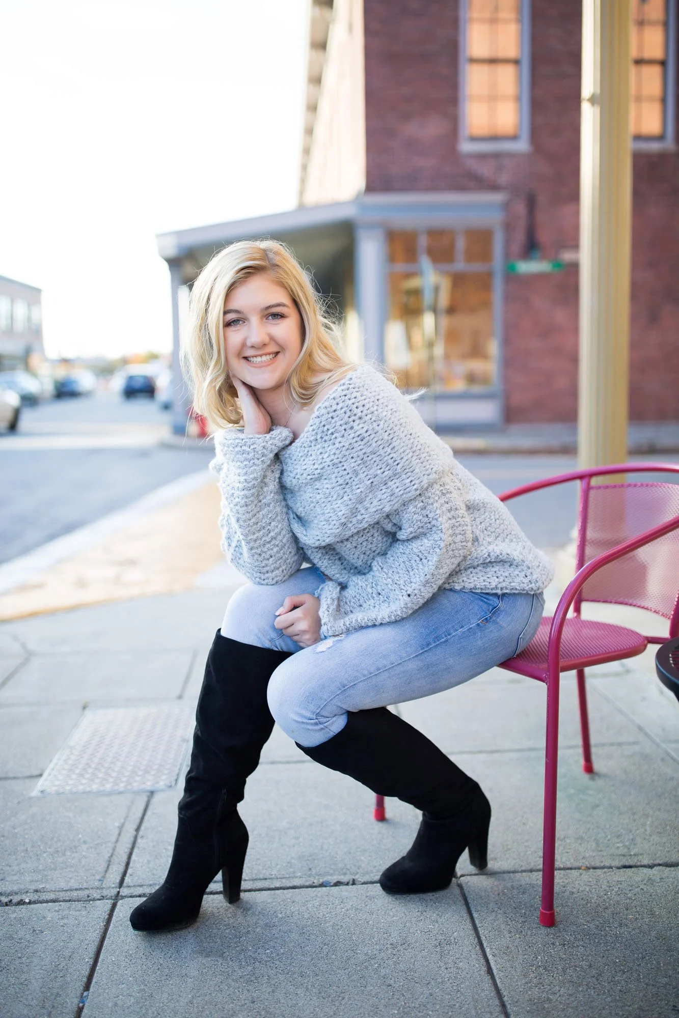 A young woman with blonde hair smiling while squatting on a sidewalk outside, wearing a gray sweater, light blue jeans, and black knee-high boots.