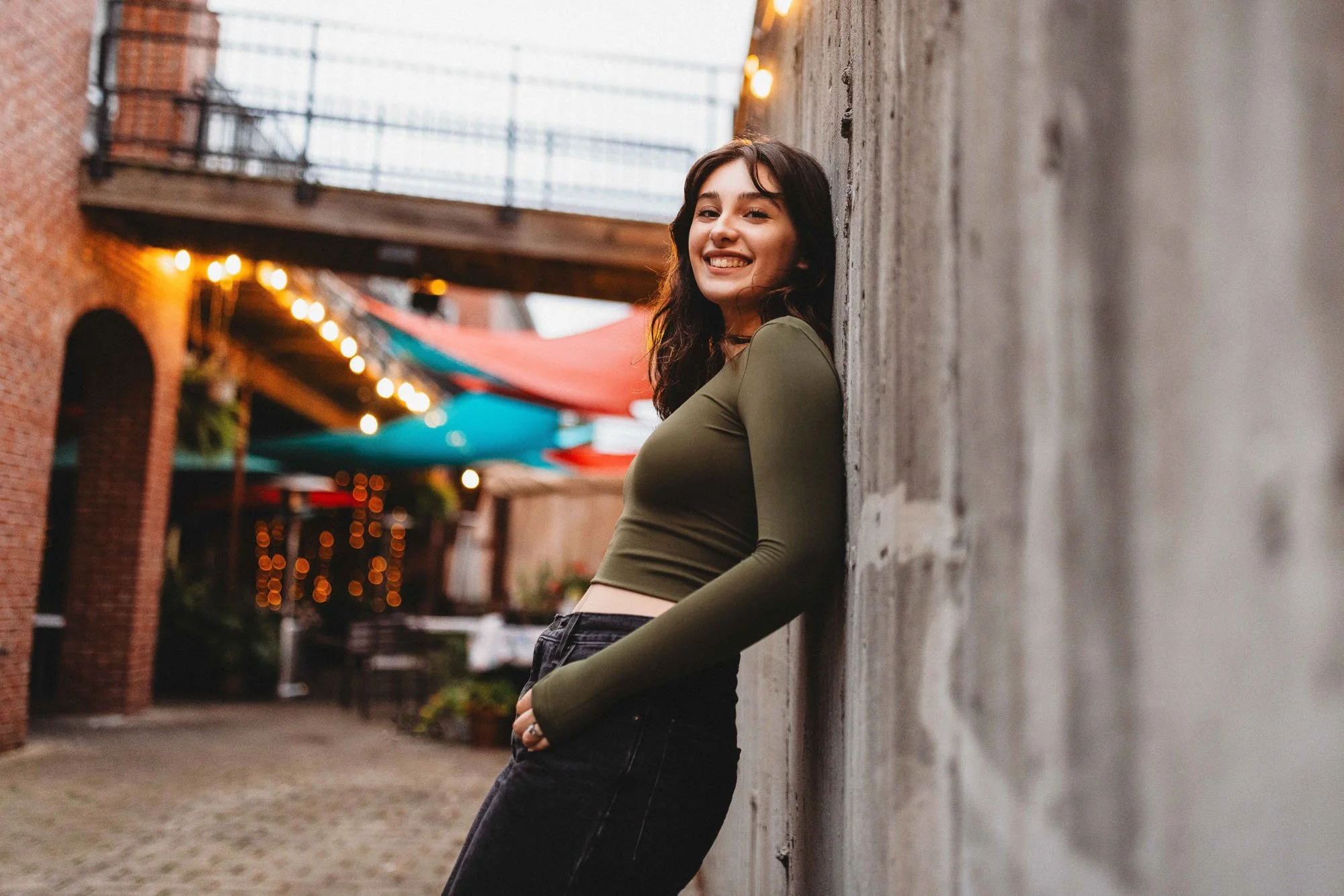Young woman with dark hair smiling, leaning against a concrete wall in an outdoor urban setting with string lights and colorful umbrellas overhead