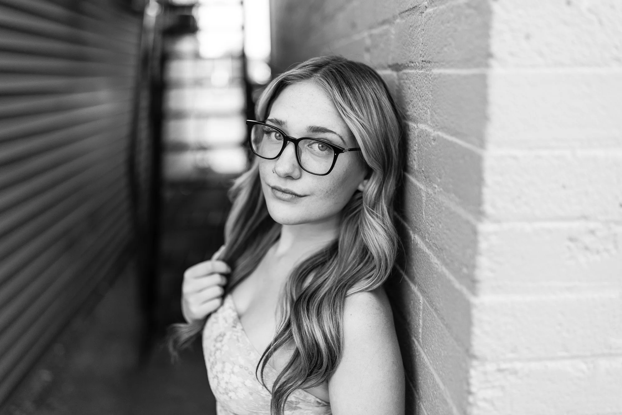 Black and white photo of a young woman with glasses and wavy hair, leaning against a brick wall in an alleyway