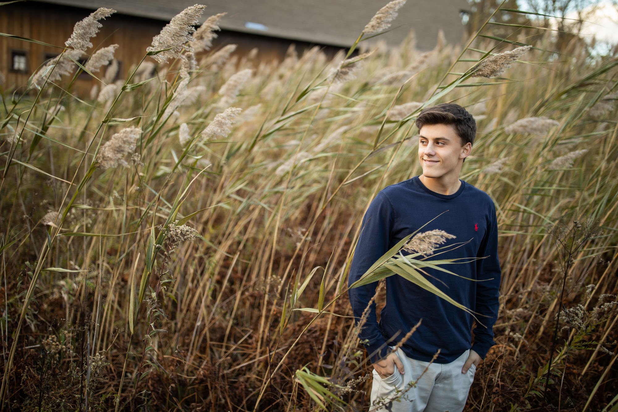 A young man with dark hair and light skin standing among tall, dry grass near a rustic wooden building. He is wearing a navy blue long-sleeve shirt and light-colored pants, with a slight smile and hands in pockets, looking to his right.