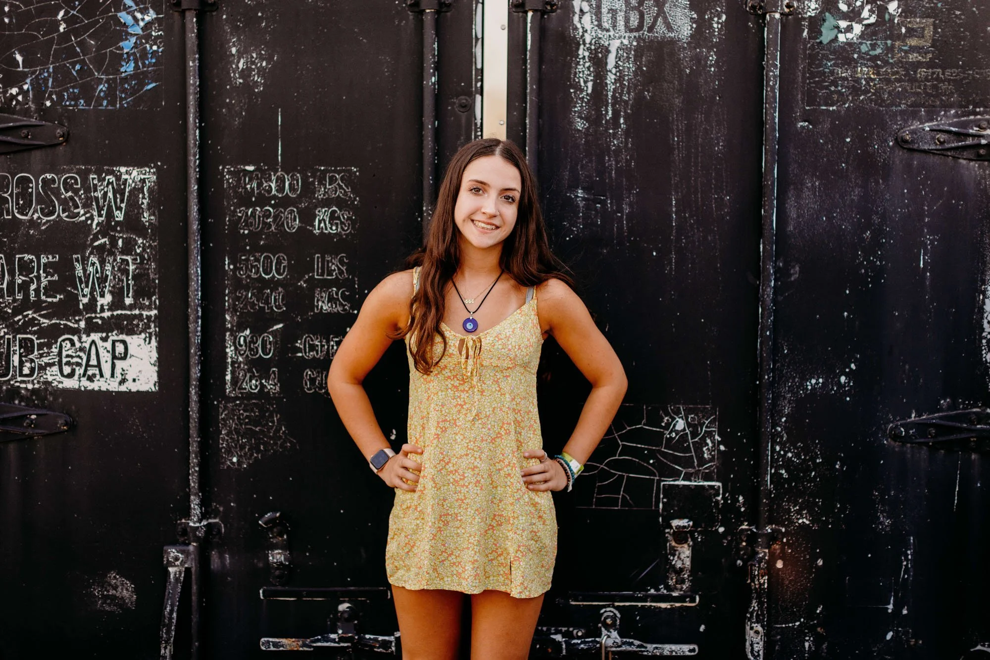 A young woman in a yellow floral dress standing with hands on hips in front of a black, graffiti-covered metal door.