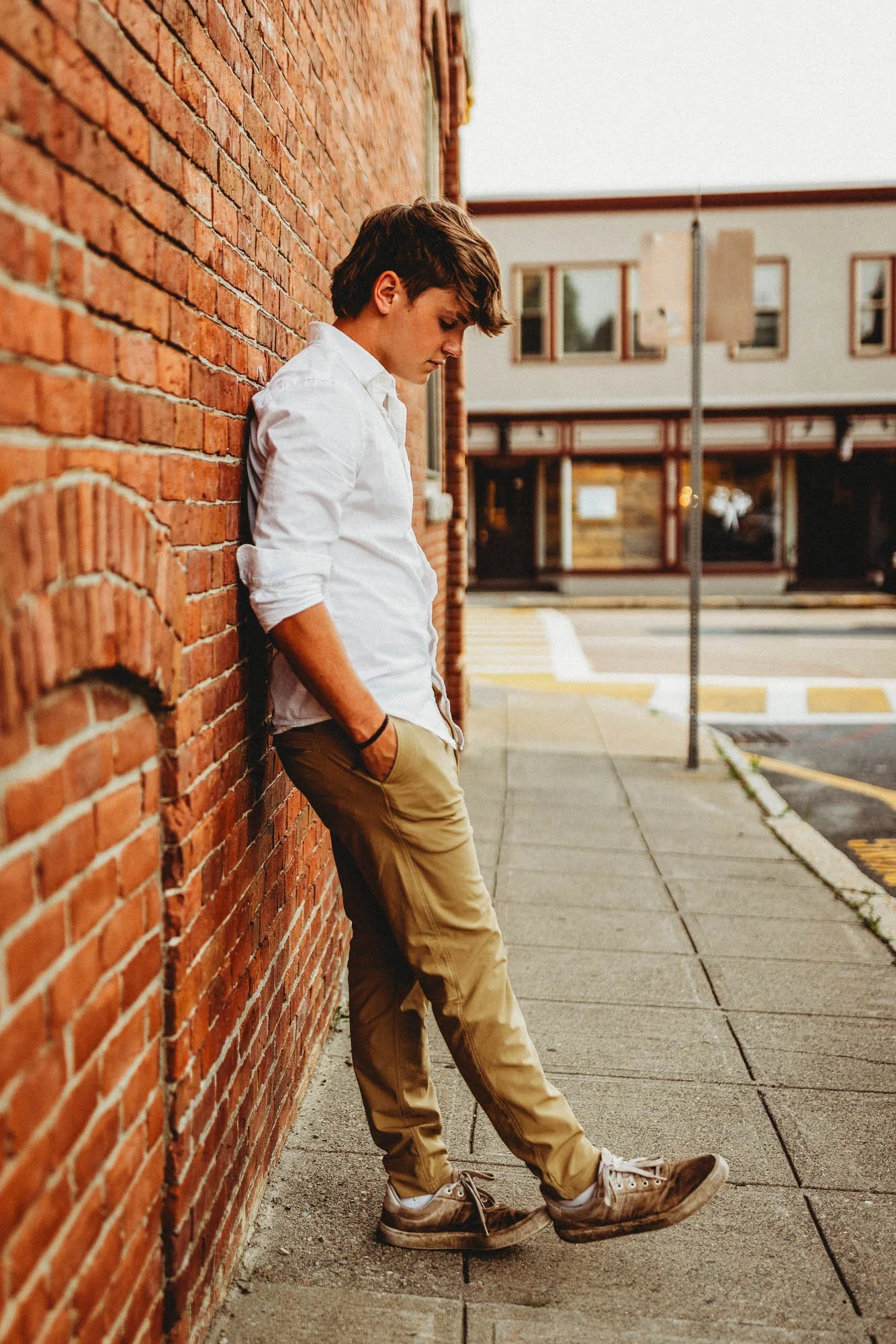 A young man with brown hair wearing a white shirt, khaki pants, and sneakers, leaning against a brick wall with his head bowed and hands in his pockets on a city sidewalk.