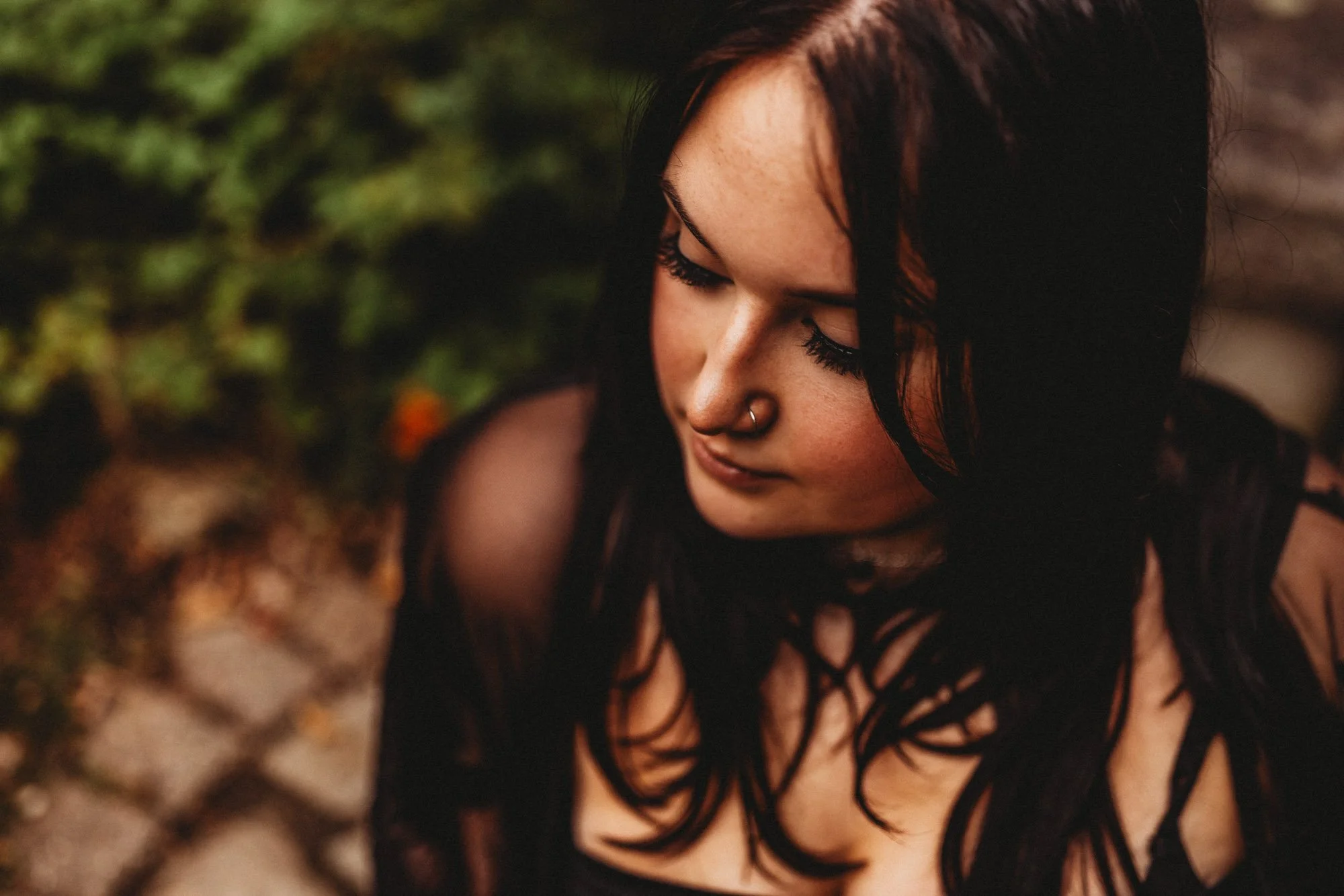 A young woman with black hair, a nose ring, and dark makeup looks downward against a blurred natural background.
