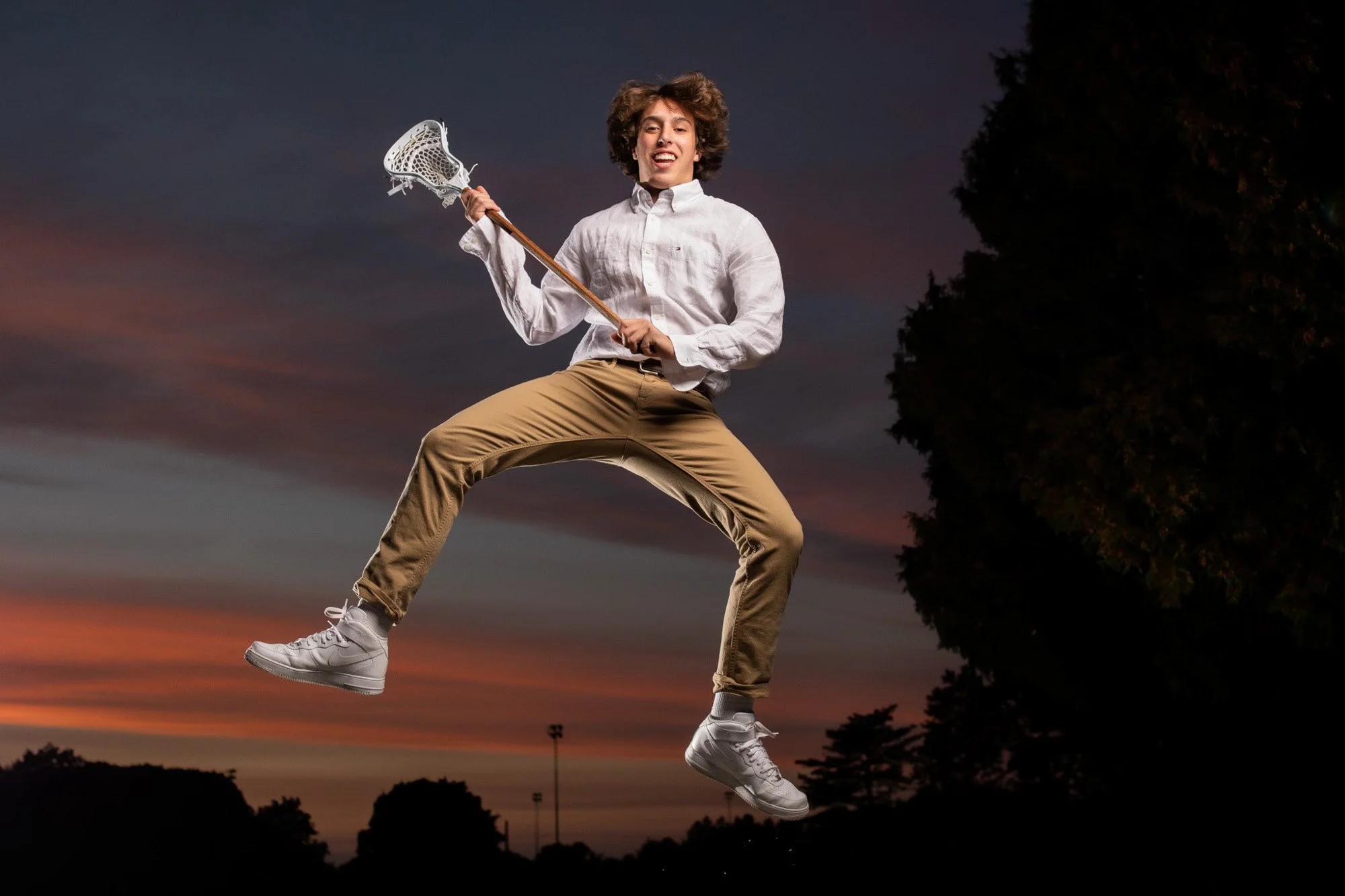 Young man jumping holding a lacrosse stick against a sunset sky and trees.