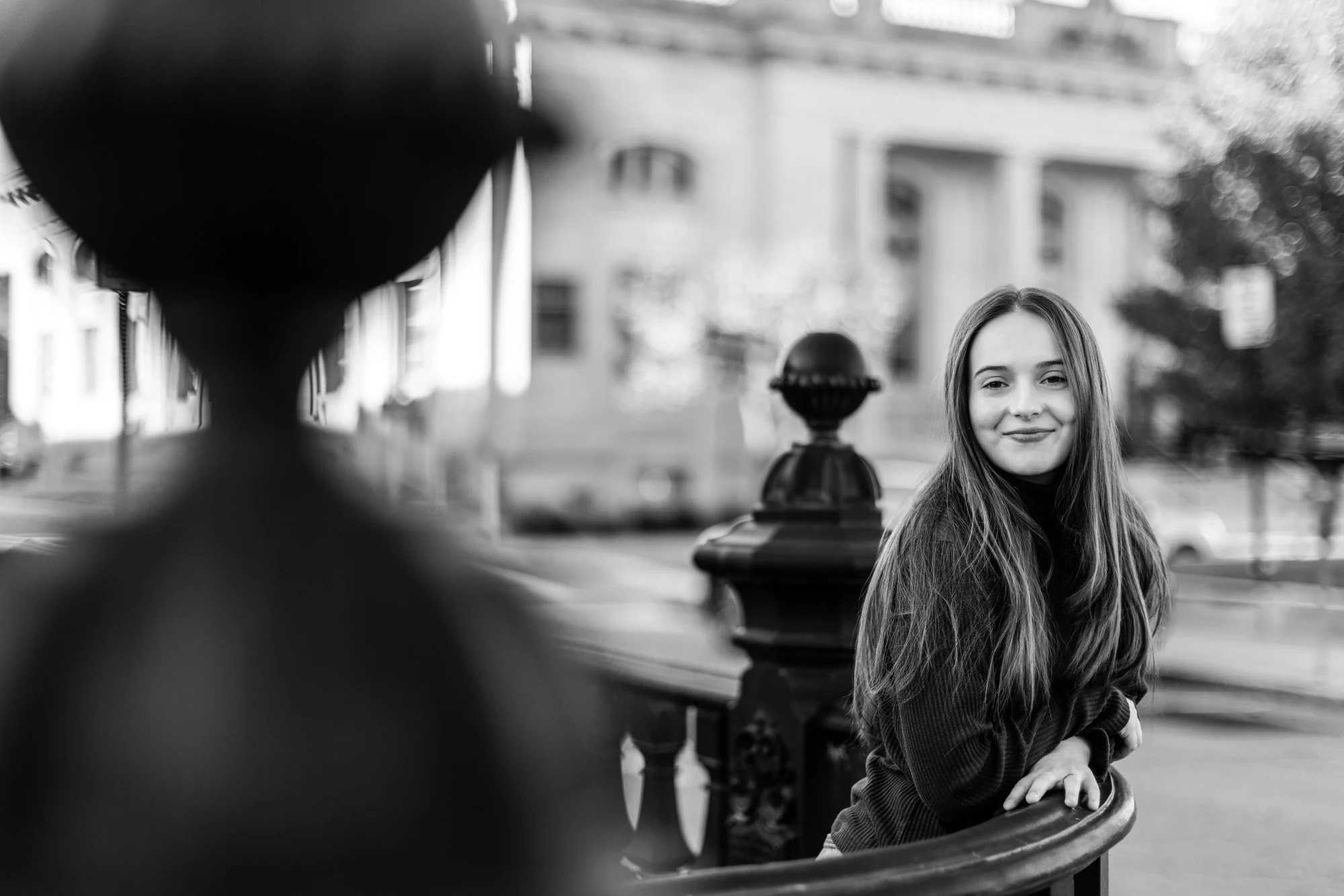 A black-and-white photo of a young woman with long hair smiling, leaning on a decorative railing outdoors, with buildings and trees in the background.