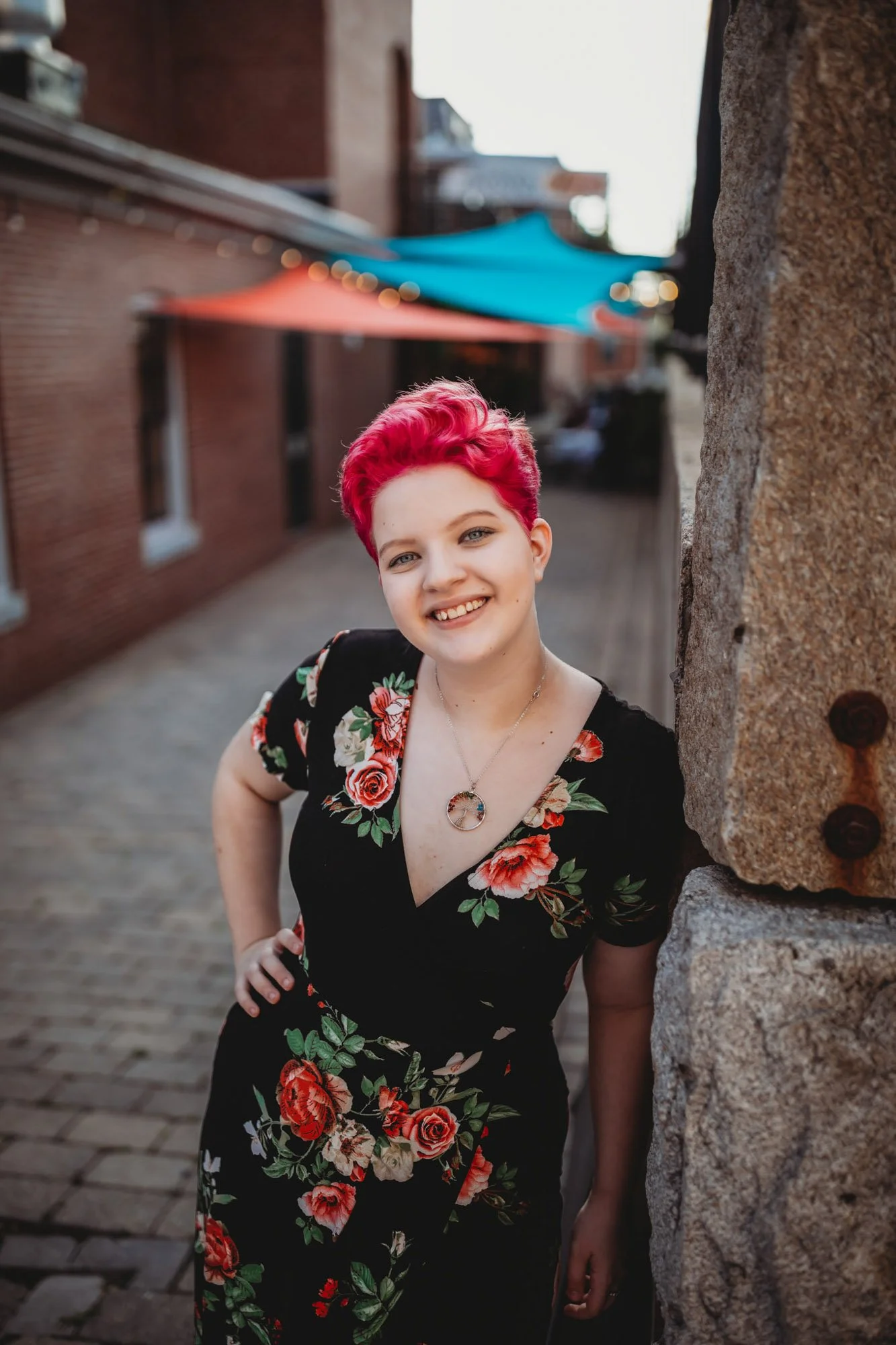A young woman with short, bright pink hair smiling and leaning against a stone wall on a cobblestone street, wearing a black dress with red and pink floral patterns and a pendant necklace.
