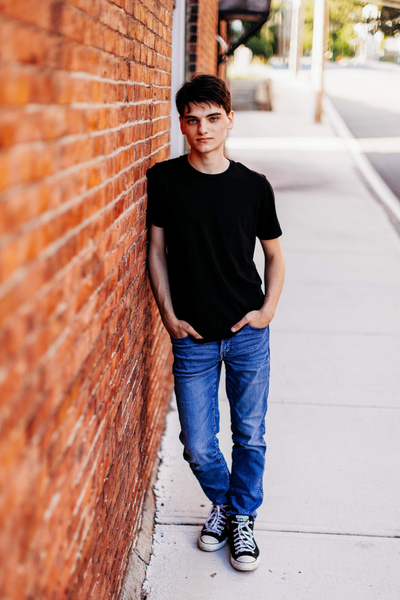 A young man with dark hair wearing a black T-shirt, blue jeans, and black sneakers standing on a sidewalk next to a brick building, with hands in pockets, looking at the camera.
