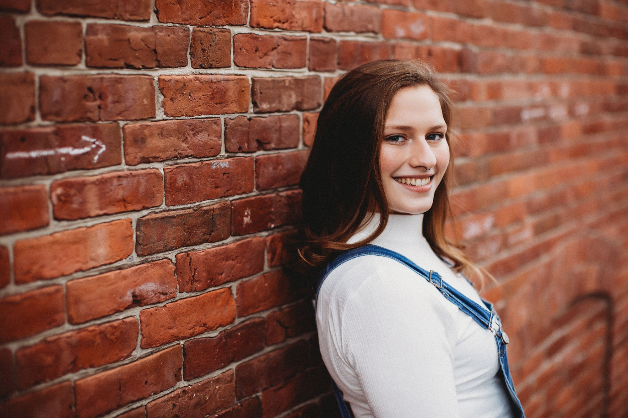 Smiling woman leaning against a red brick wall, wearing a white turtleneck and blue overalls.