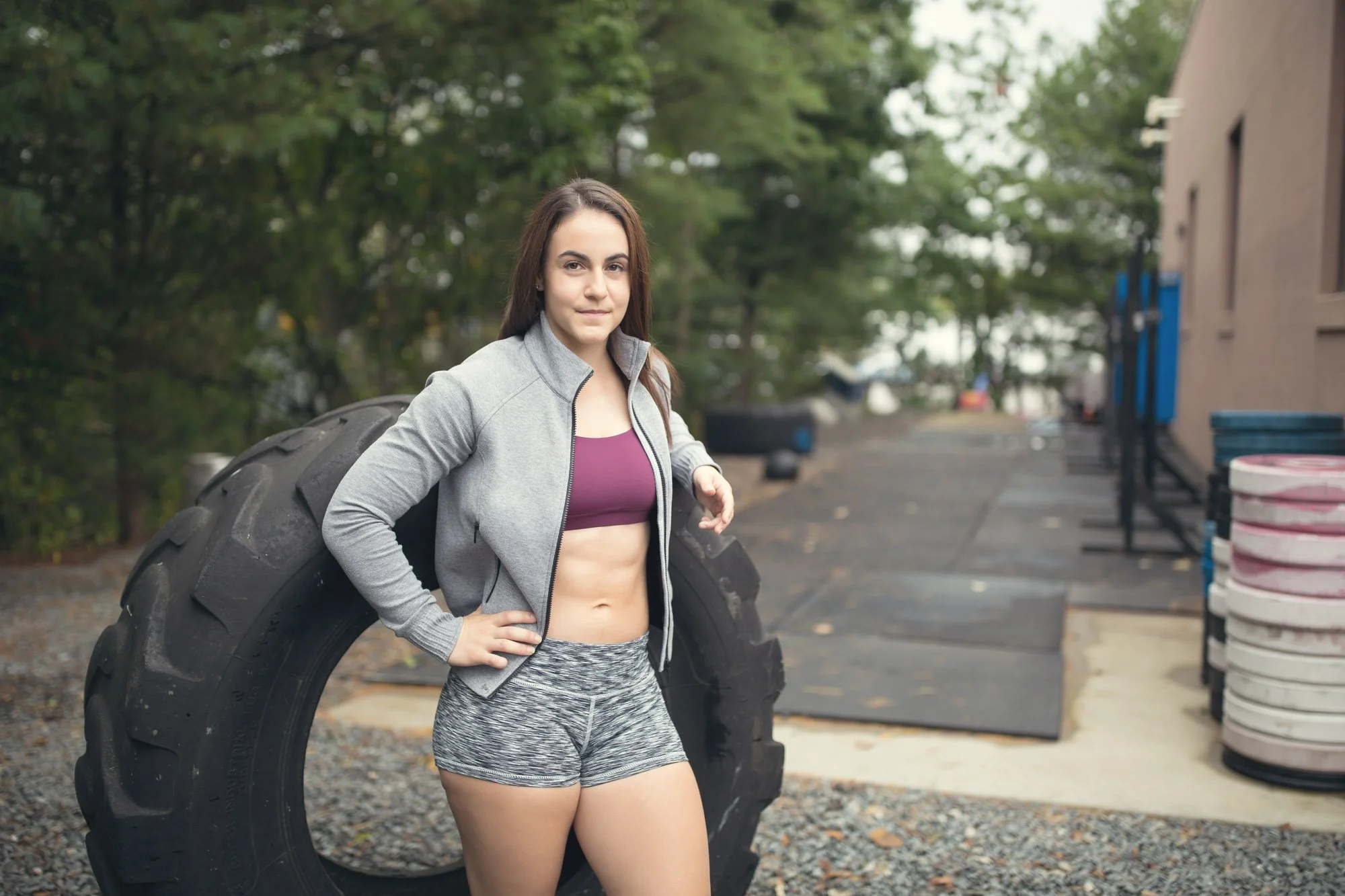 Young woman in workout clothes standing outdoors with one hand on her hip next to a large tire.