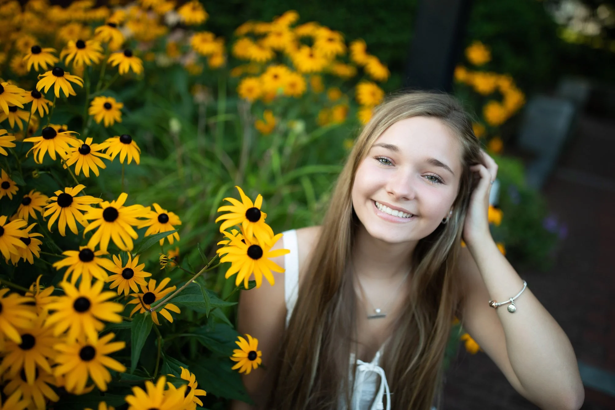 A young woman with long blonde hair smiling at the camera while surrounded by yellow and black flowers in a garden.