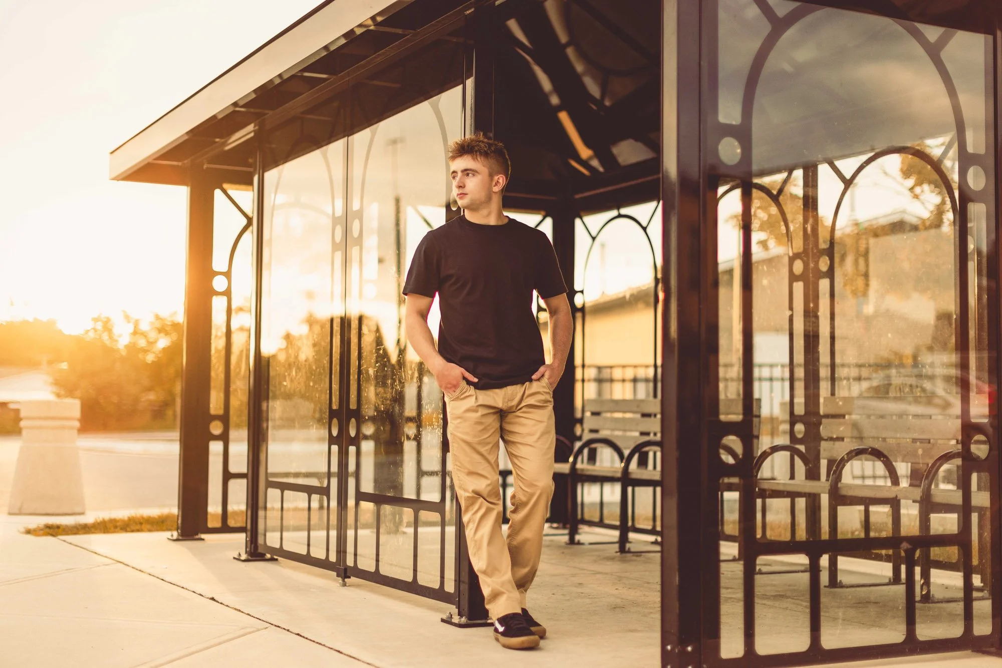 Young man in a black t-shirt and beige pants standing inside a bus stop shelter at sunset.