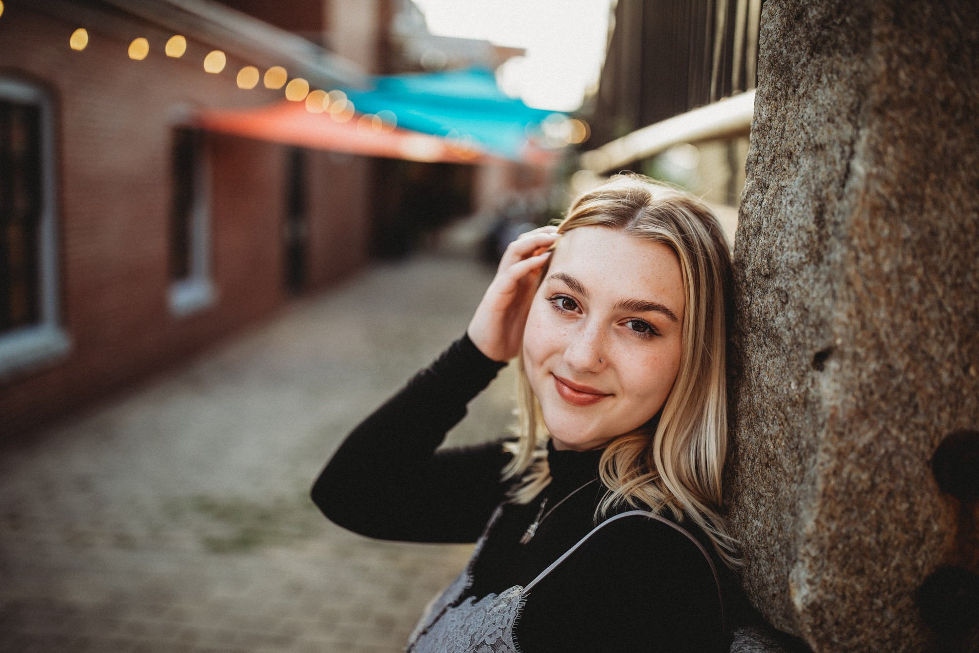 A young woman with blonde hair and a black top, smiling and leaning her head against a stone wall on a street, with blurred buildings and string lights in the background.