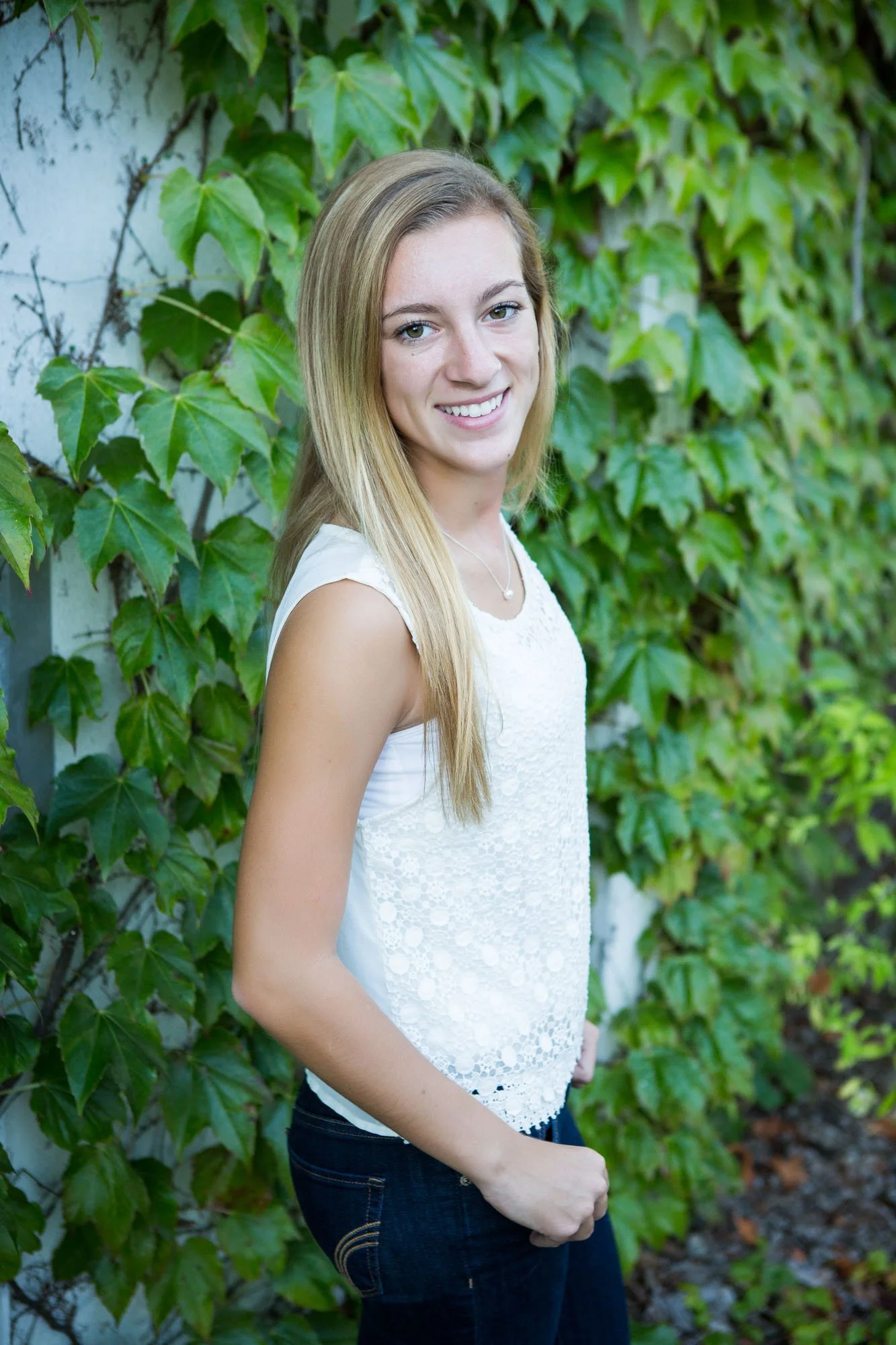 A young woman with long blonde hair smiling outdoors, standing in front of green ivy-covered wall, wearing a white sleeveless lace top and dark jeans.