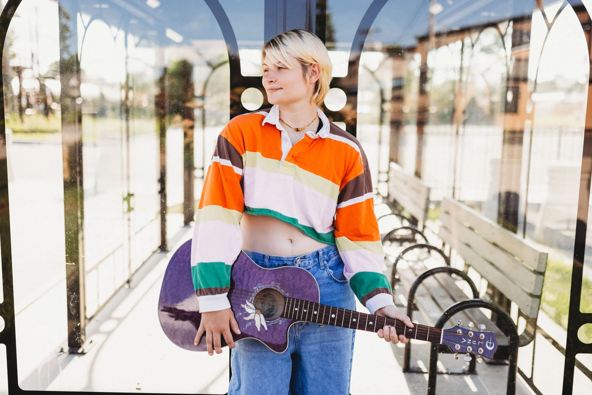 A young woman with short blonde hair standing at a bus stop, holding a purple acoustic guitar with a unicorn design, wearing a colorful striped cropped jacket and blue jeans.