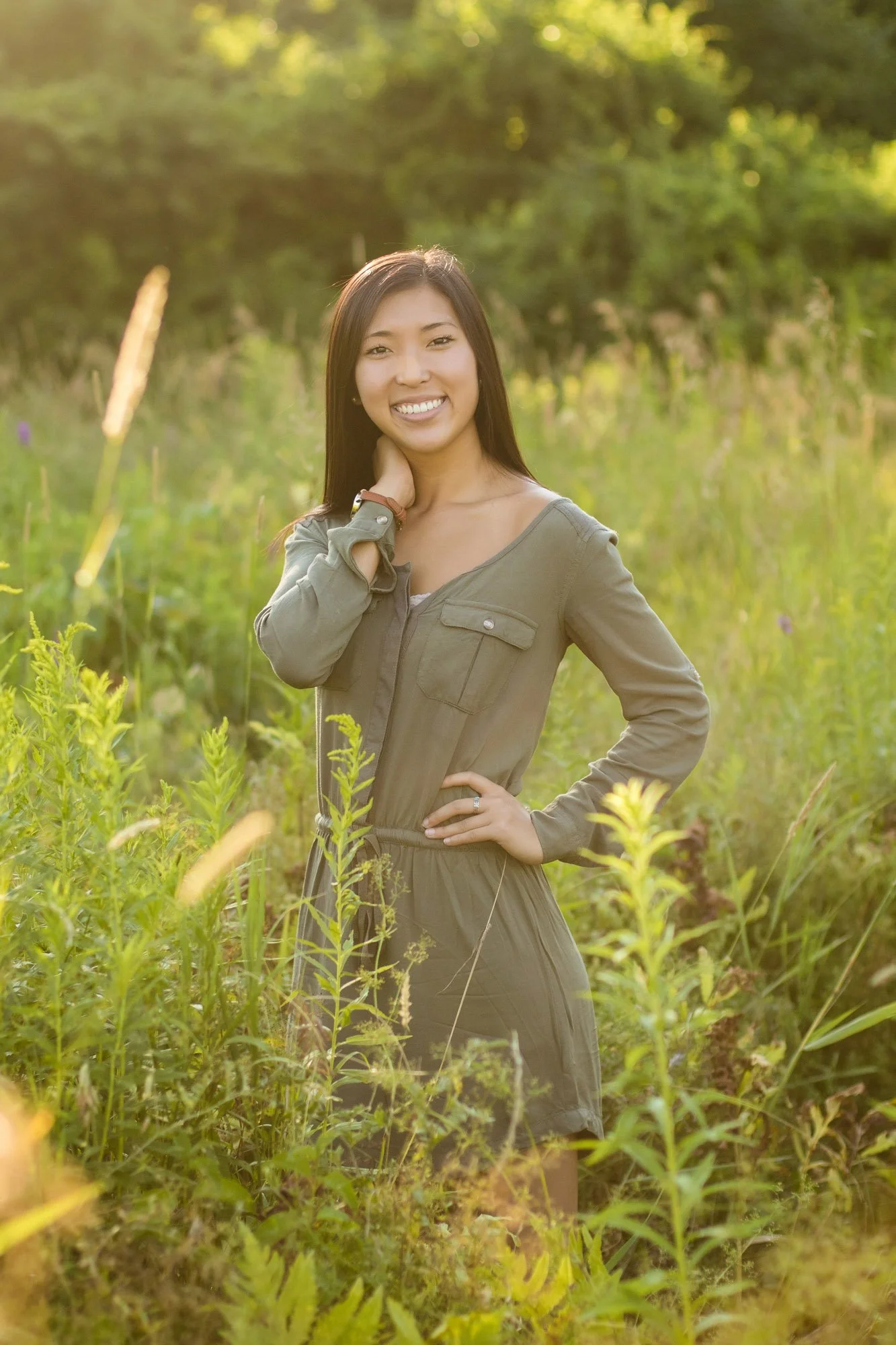 Young woman in a green jumpsuit standing in a grassy field with trees in the background, smiling at the camera during golden hour sunlight.