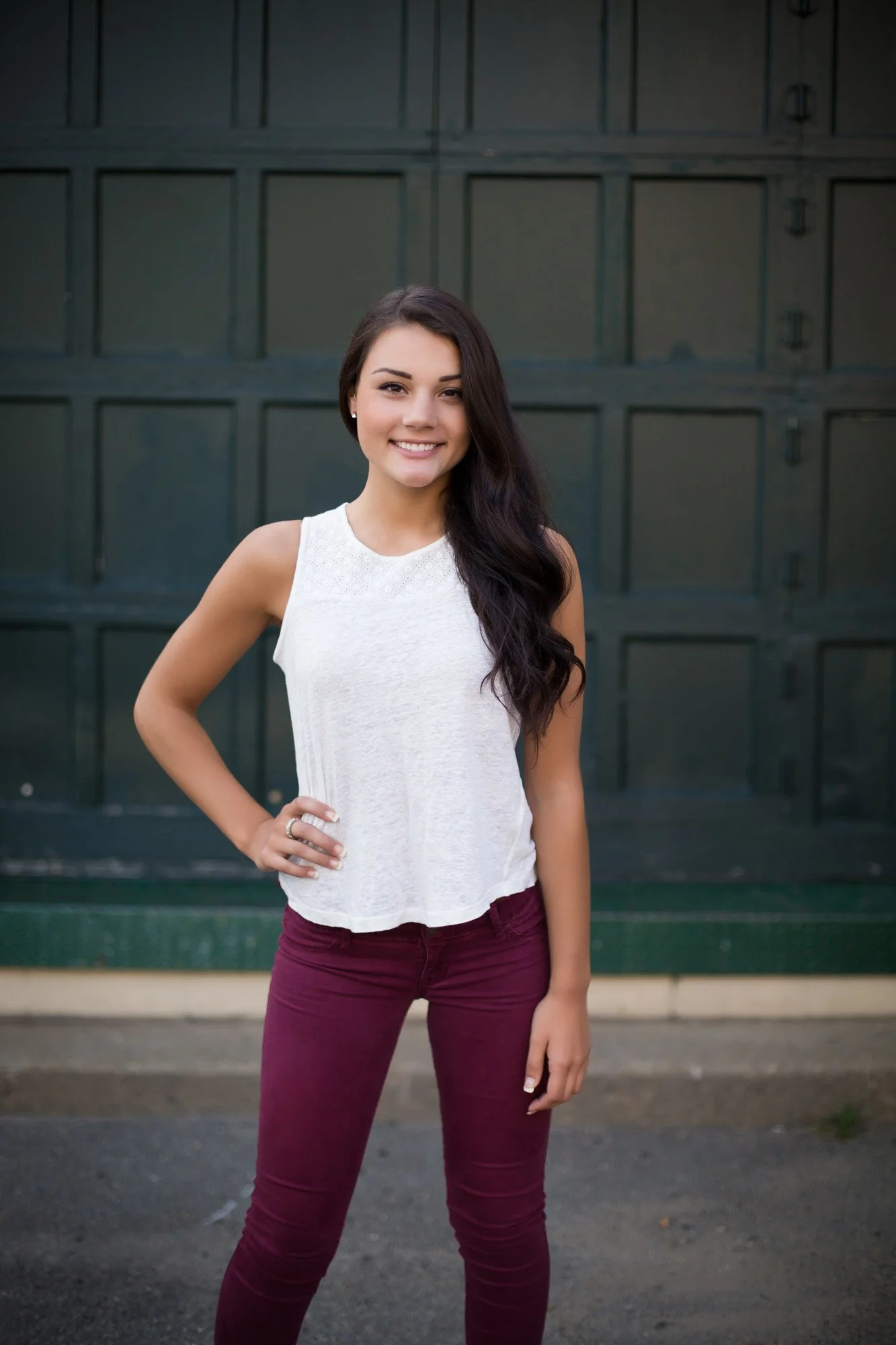 A young woman with long dark hair wearing a sleeveless white top and maroon pants, standing outside in front of a dark green garage door, smiling with her hand on her hip.