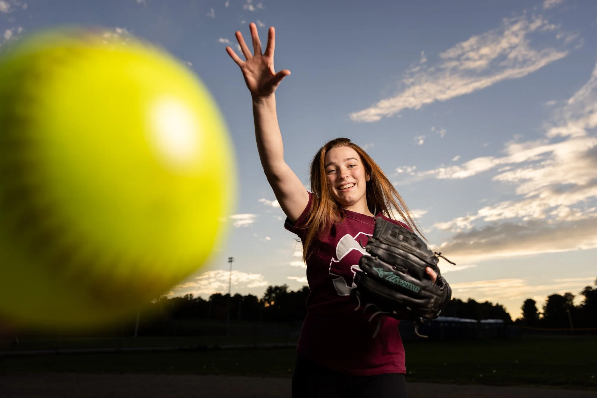 Young woman in a maroon shirt throwing a yellow softball with a baseball glove in her hand at a sports field during sunset.