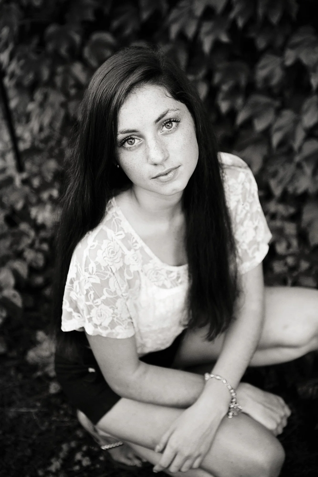 Black and white photo of a young woman with long dark hair, wearing a lace top, sitting on the ground, looking directly at the camera with a neutral expression, row of leaves in the background.
