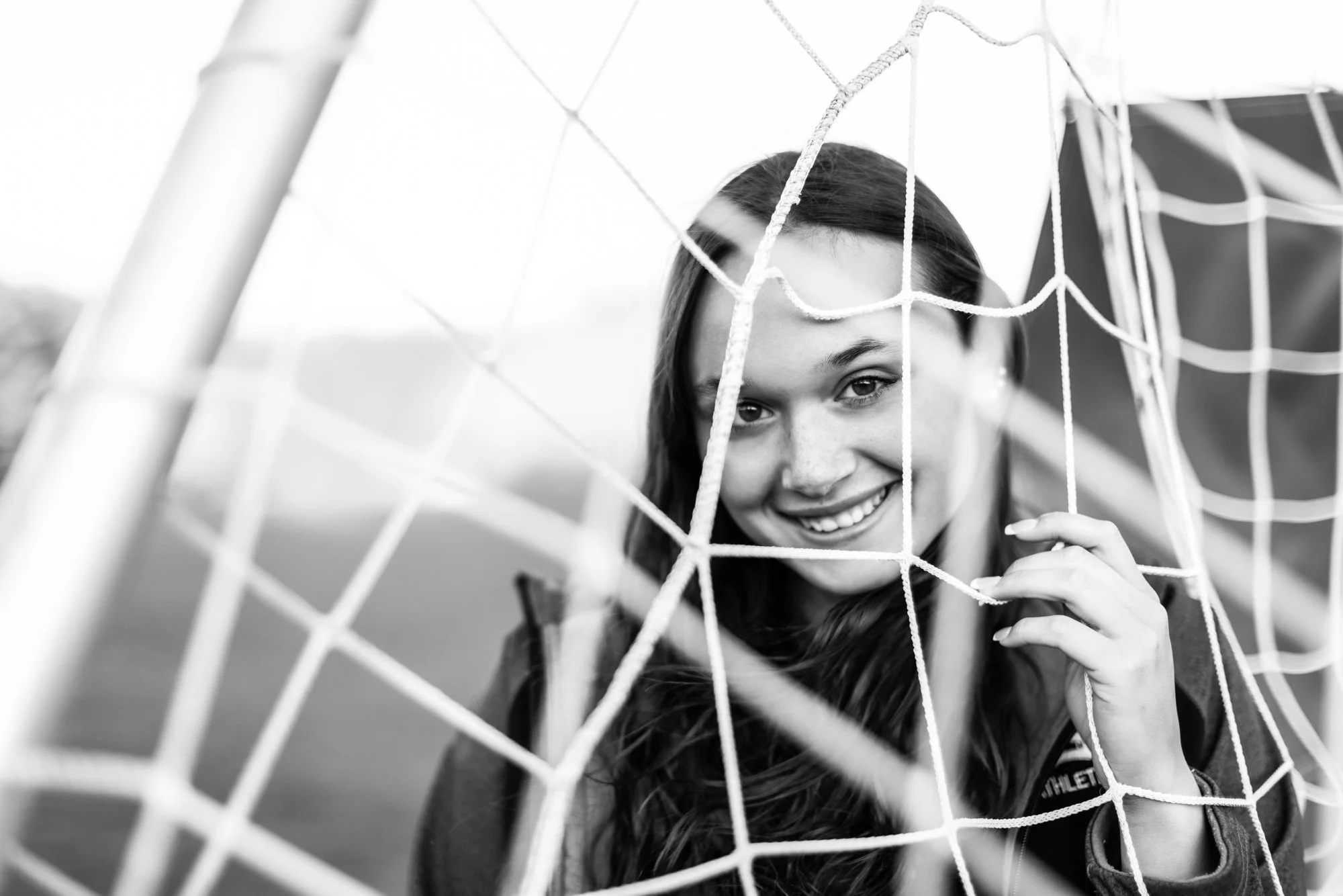 Black and white photo of a smiling young woman with long hair behind a soccer goal net, looking through it.