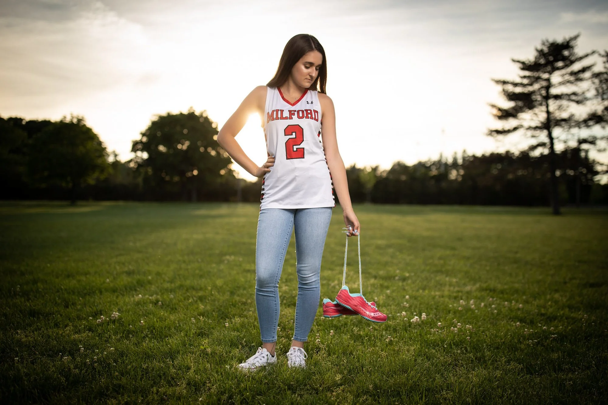 A young woman in a Milford basketball jersey and jeans stands on a grassy field, holding a pair of pink running shoes on a string, with trees and a sunset background.