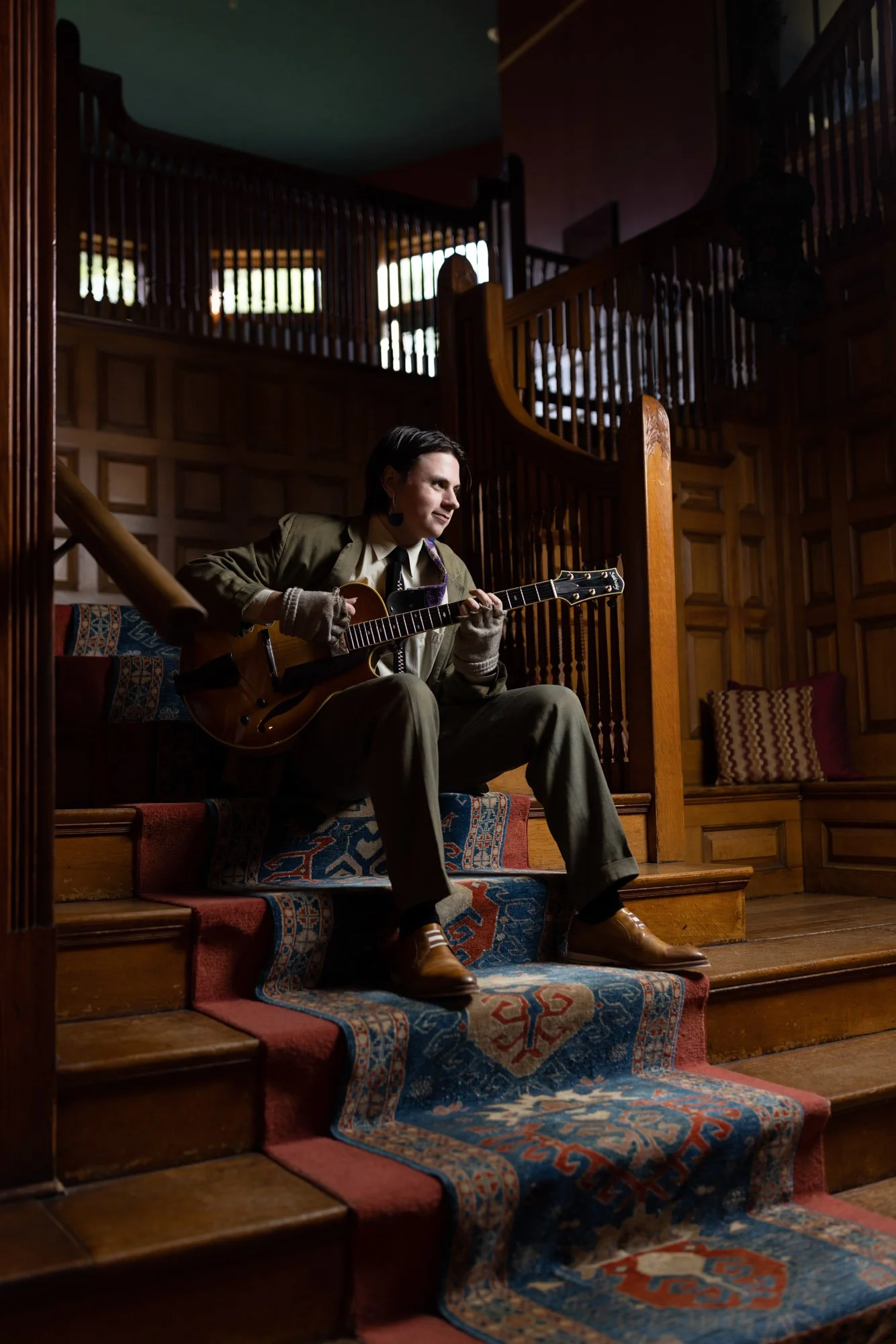 A person wearing a dark jacket, light shirt, and gloves sitting on a staircase playing an electric guitar, with a wooden interior background.
