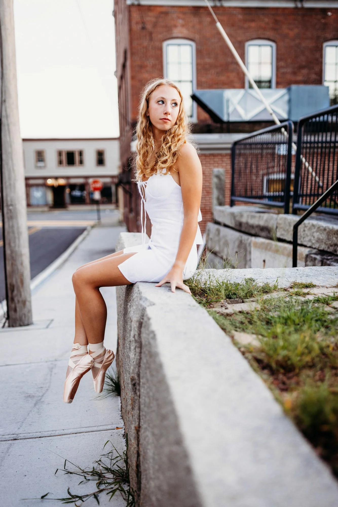 A young woman with curly blonde hair, wearing a white dress and ballet shoes, sits on a concrete ledge on a city sidewalk.