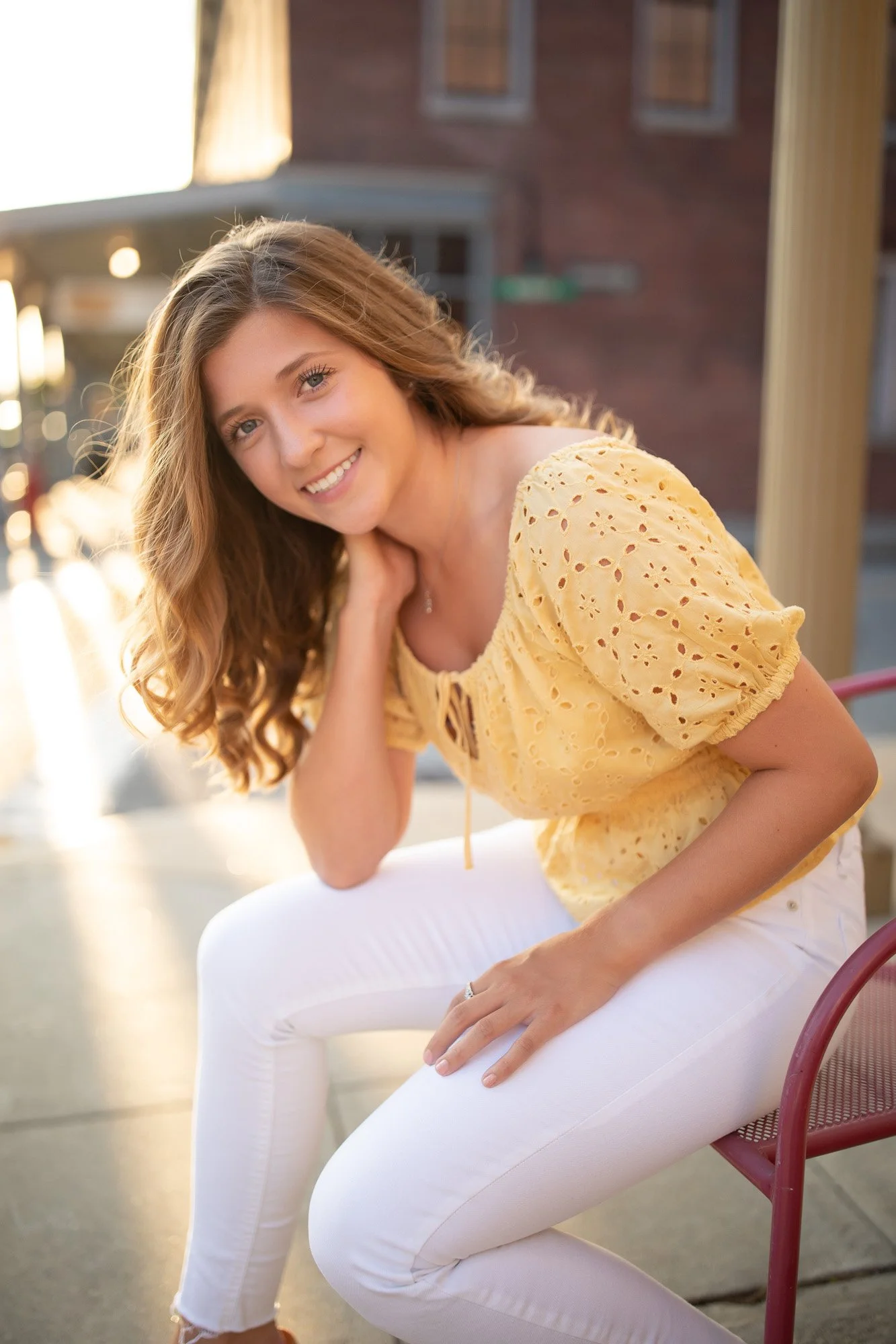 A young woman with wavy, shoulder-length hair sitting on a red chair outdoors, wearing a yellow eyelet blouse and white pants, smiling at the camera.