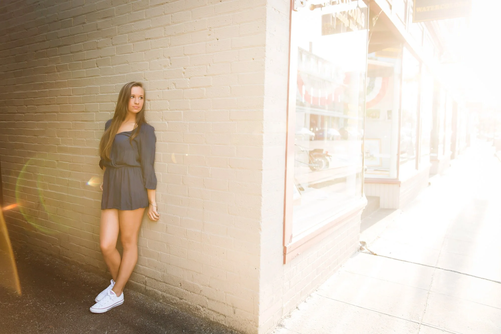A young woman with long brown hair, wearing a black dress and white sneakers, standing against a beige brick wall on a sidewalk, with sunlight causing lens flare in front of a storefront window.