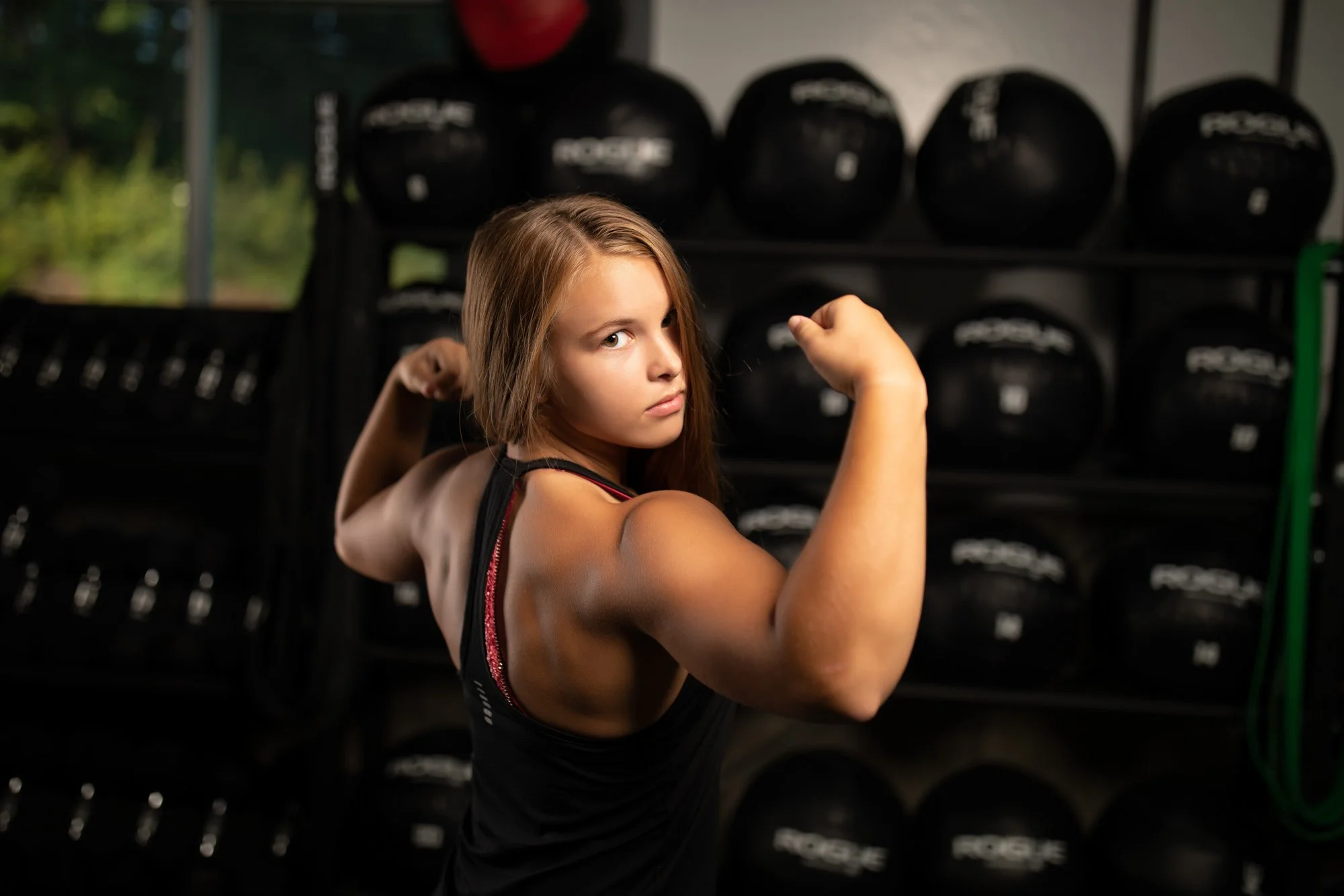 A young woman flexing her arms in a gym with black medicine balls in the background.