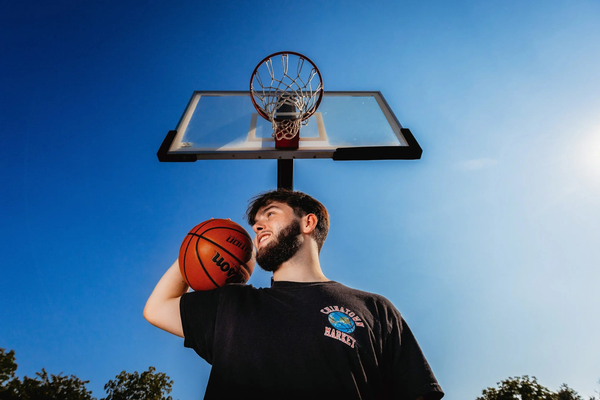 A young man with a beard holding a basketball, standing under a basketball hoop against a bright blue sky, smiling and looking away.