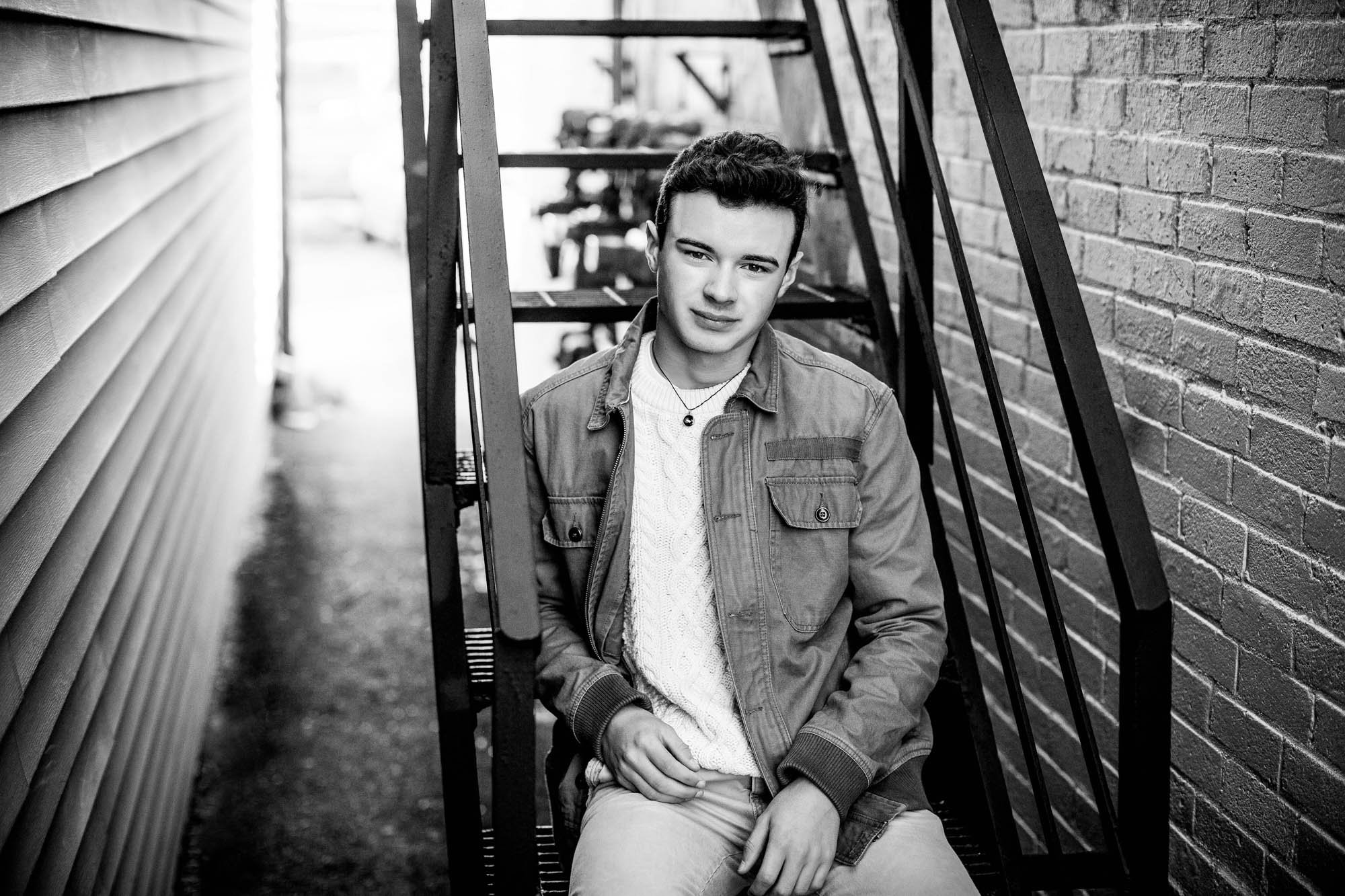 Black and white photo of a young man sitting on a fire escape between a brick wall and siding, looking at the camera.