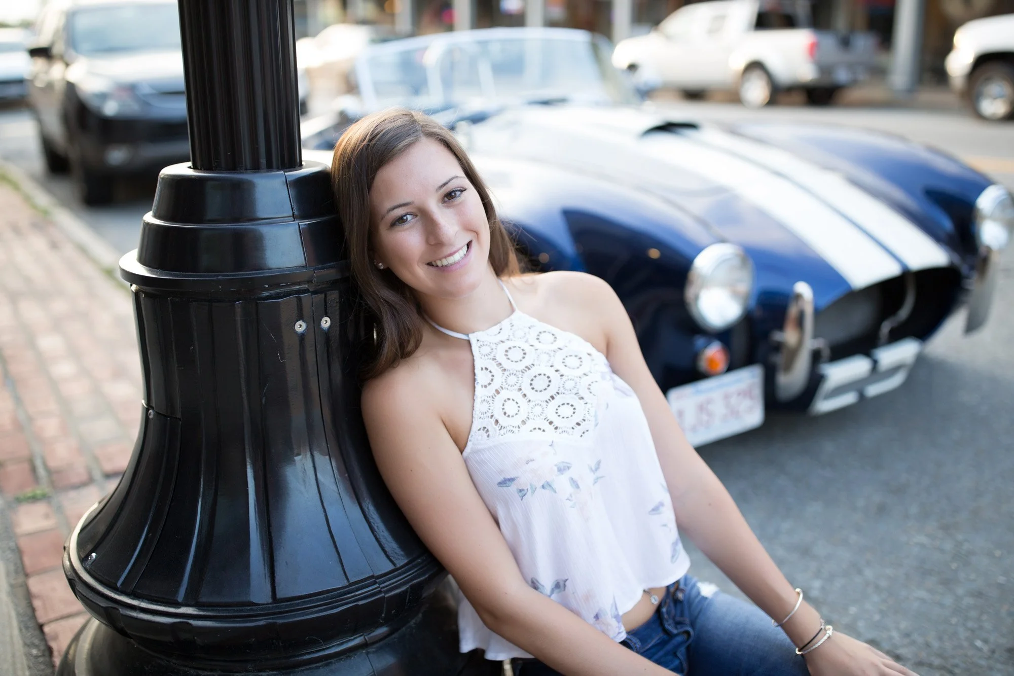 A young woman with brown hair sitting and smiling by a black street lamp, with a vintage blue and white race car in the background.