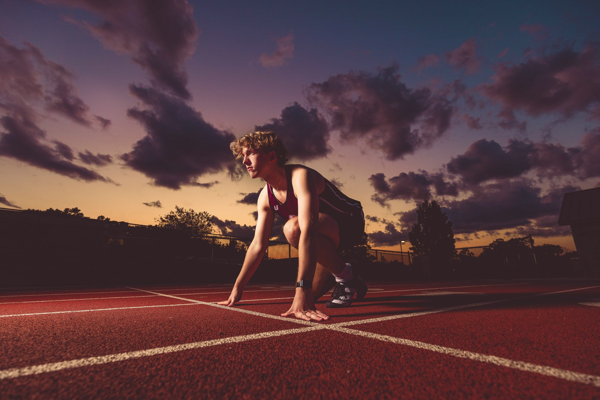A young male athlete in a starting position on a running track during sunset, with clouds in the sky.