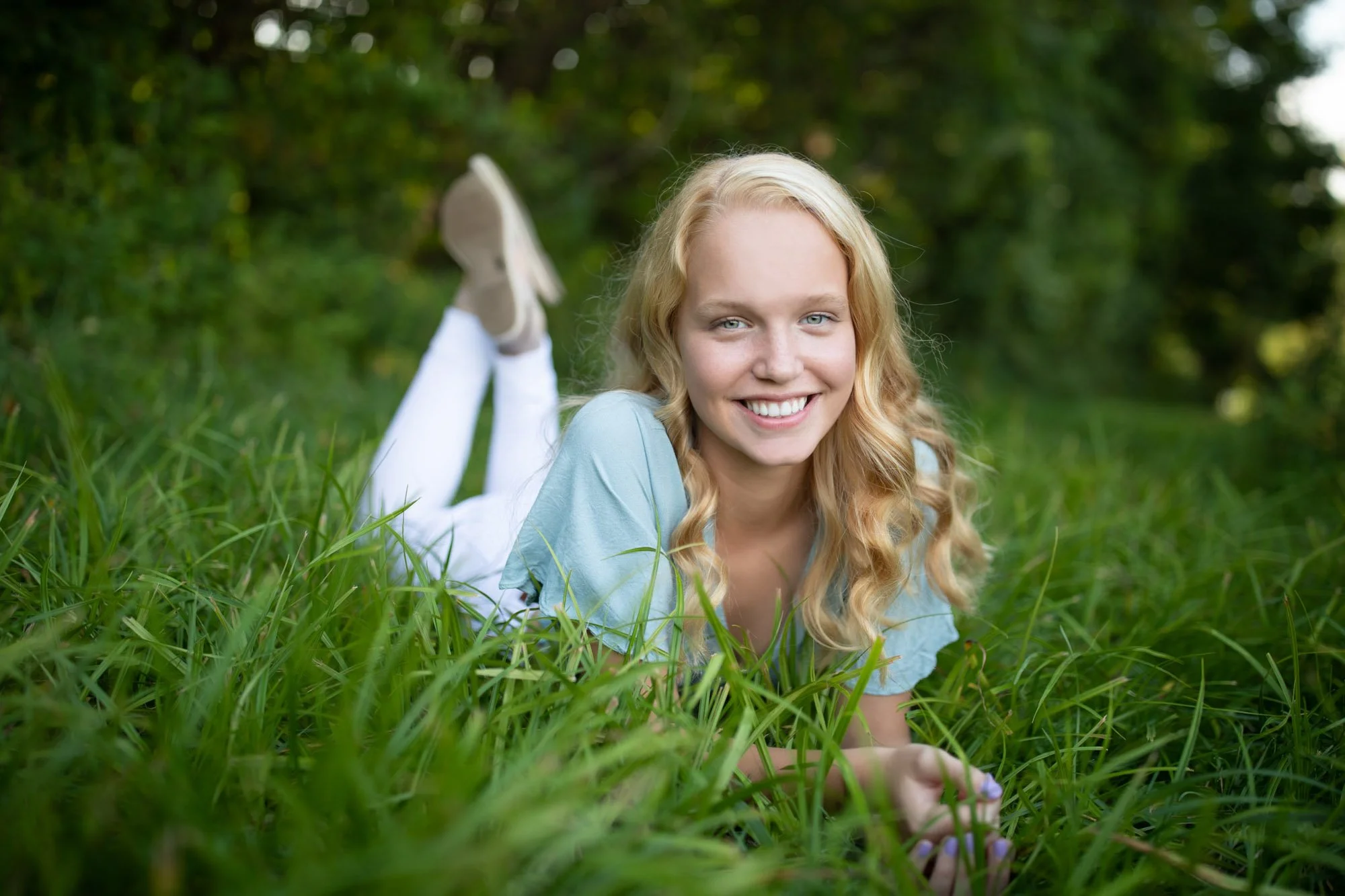 Young woman with long blonde curly hair lying on her stomach in tall green grass outdoors, smiling at the camera, with trees and greenery in the background.